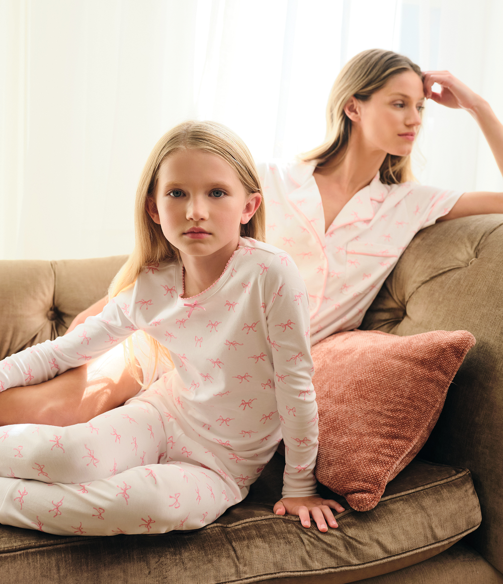 A young girl in white pajamas with pink patterns sits on a couch next to a woman in matching pajama top and pants. The woman leans back on a brown cushion, looking away, while sunlight streams in through sheer curtains.
