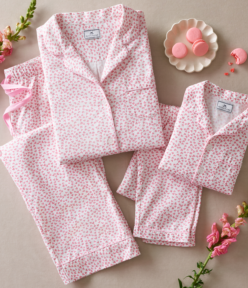 Two sets of matching pink and white heart-patterned pajamas, one adult and one child-sized, are neatly folded on a beige surface with pink flowers and a plate of pink macarons beside them.