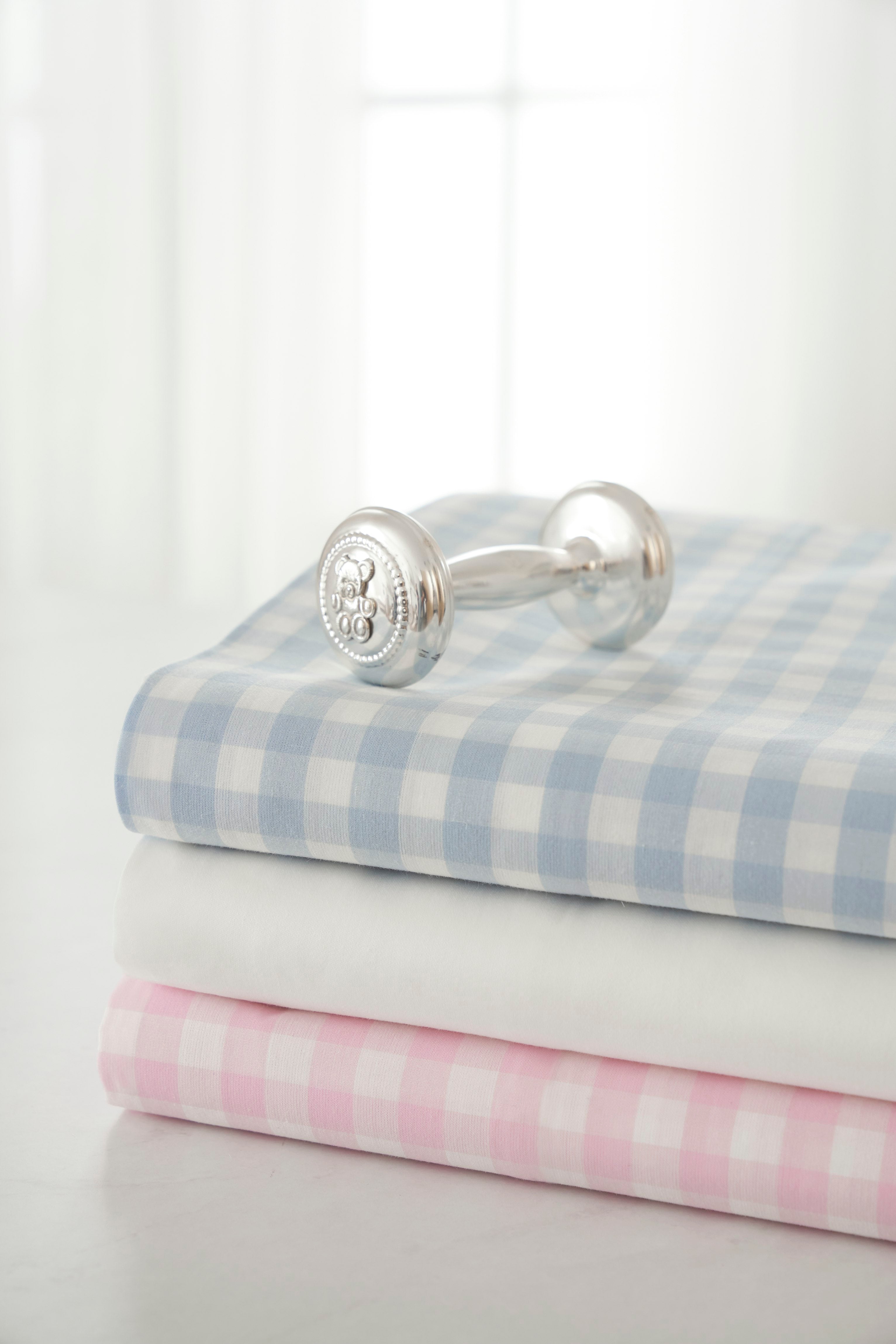 A silver baby rattle sits on three folded fabrics, one being the Luxe Premium Cotton Pink Gingham Crib Sheet by Petite Plume, all stacked on a table. A softly lit window with sheer curtains in the background enhances the blue, white, and pink checkered pattern, creating a bright and serene atmosphere.