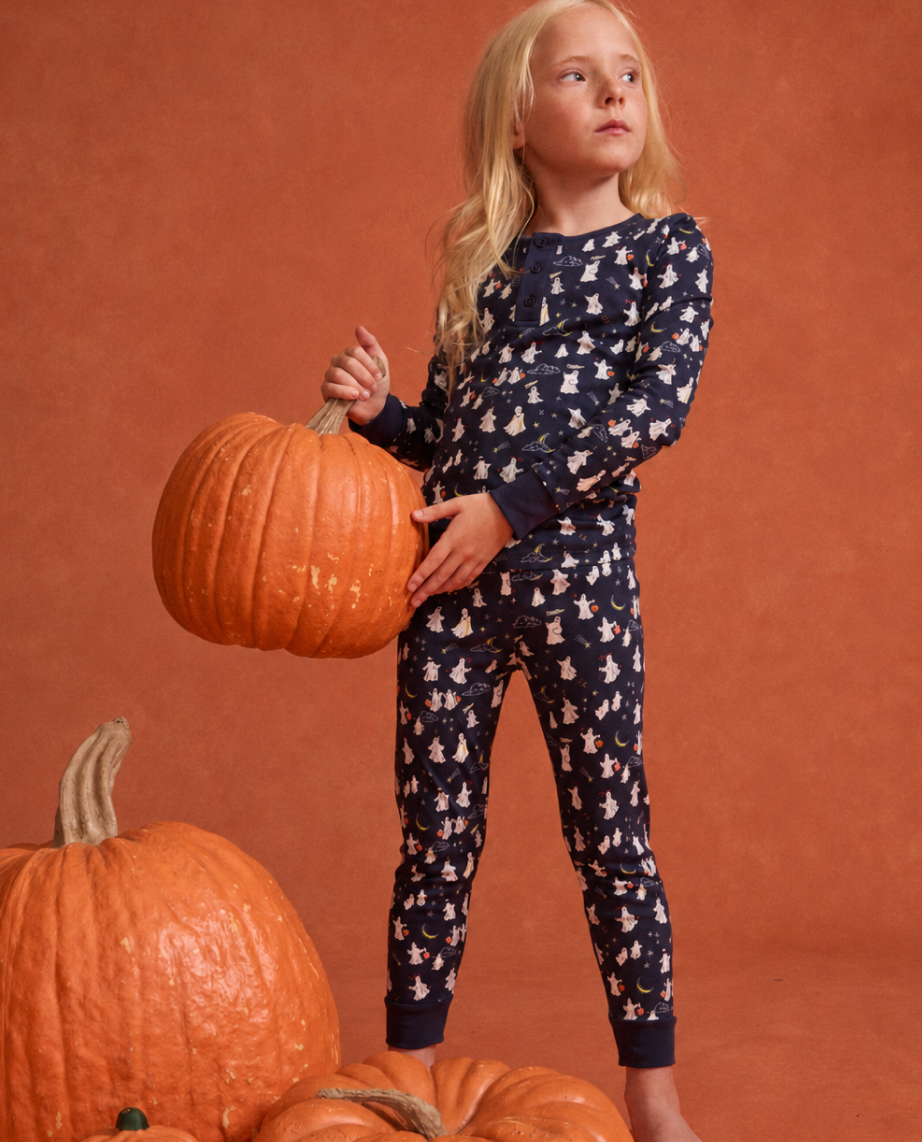 A child in navy pajamas with white and orange prints holds a large pumpkin, standing among more pumpkins on an orange background, looking off to the side.
