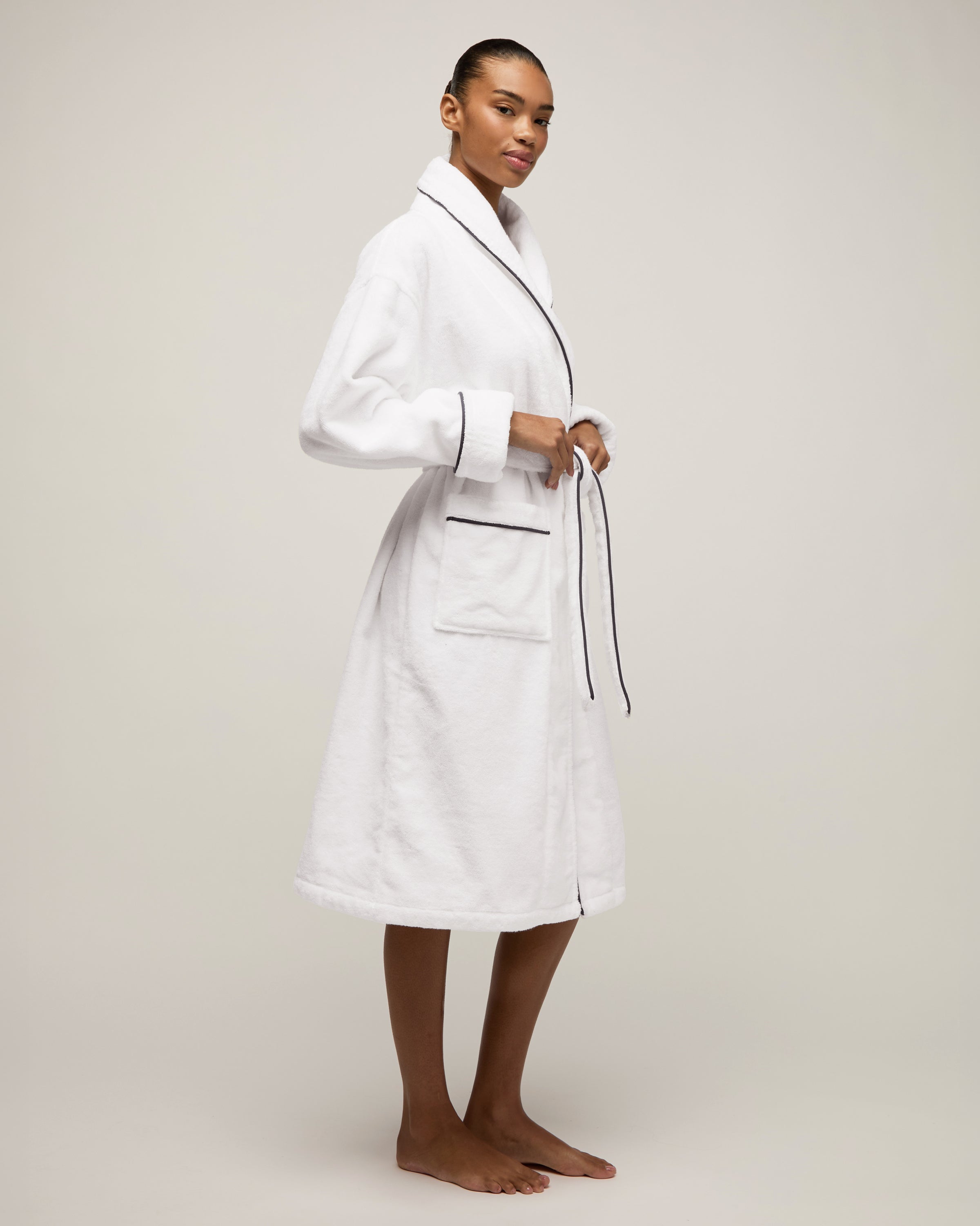 The Unisex Luxe Terry Robe in White with Navy Piping has a shawl collar, front pockets, waist tie, rolled sleeves, and dark piping along the edges.