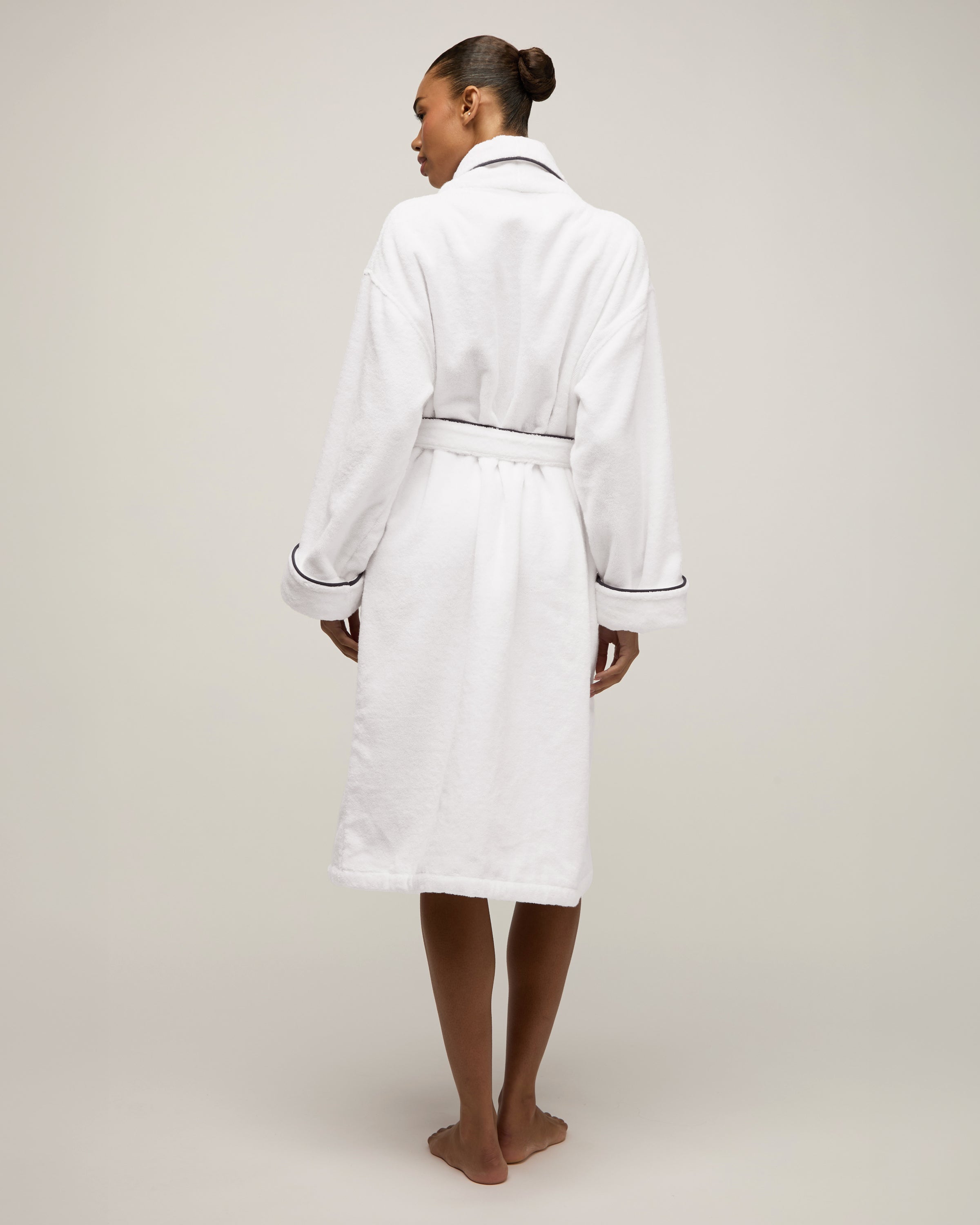 The Unisex Luxe Terry Robe in White with Navy Piping has a shawl collar, front pockets, waist tie, rolled sleeves, and dark piping along the edges.