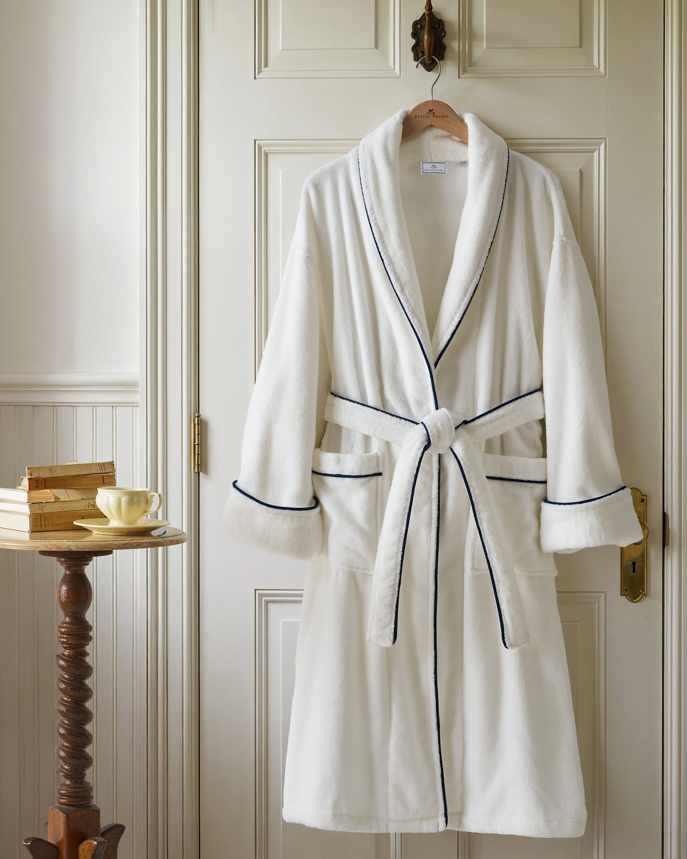 The Unisex Luxe Terry Robe in White with Navy Piping has a shawl collar, front pockets, waist tie, rolled sleeves, and dark piping along the edges.