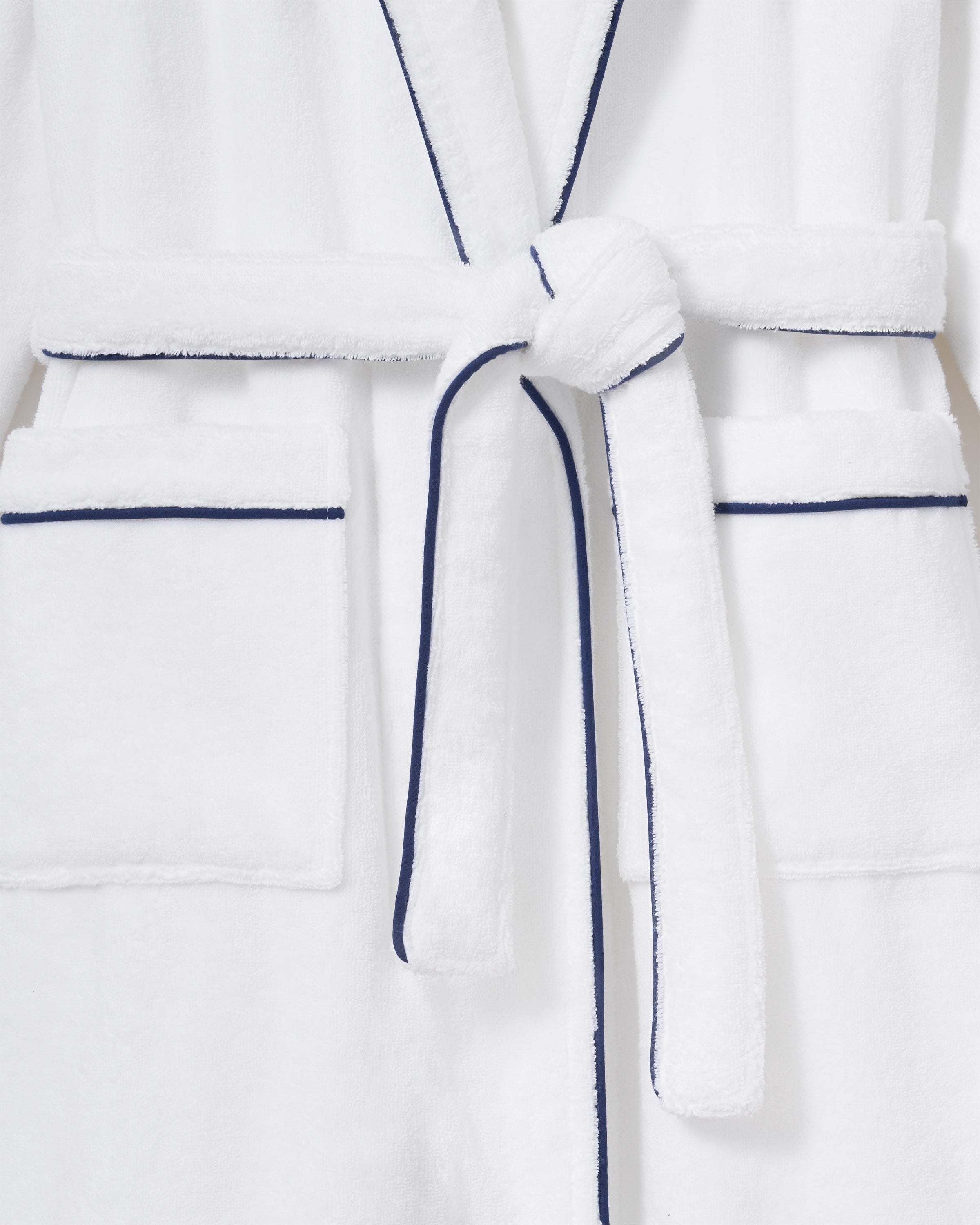 The Unisex Luxe Terry Robe in White with Navy Piping has a shawl collar, front pockets, waist tie, rolled sleeves, and dark piping along the edges.