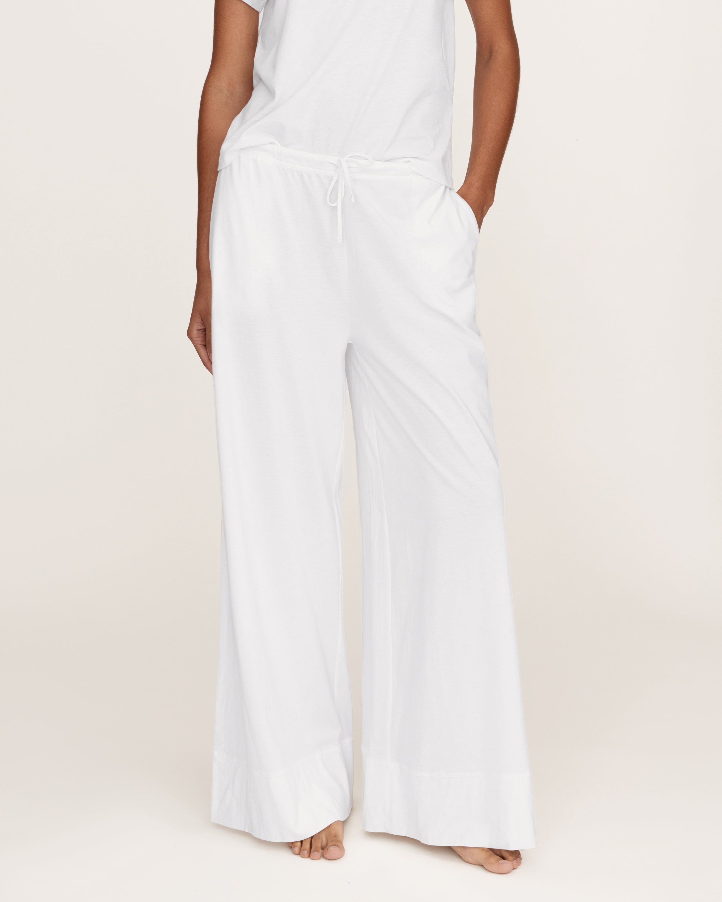 A person with long curly hair stands against a plain light background, wearing a loose-fitting white top and the Women's Pima Wide-Leg Lounge Pant in White, with one hand in a pocket.