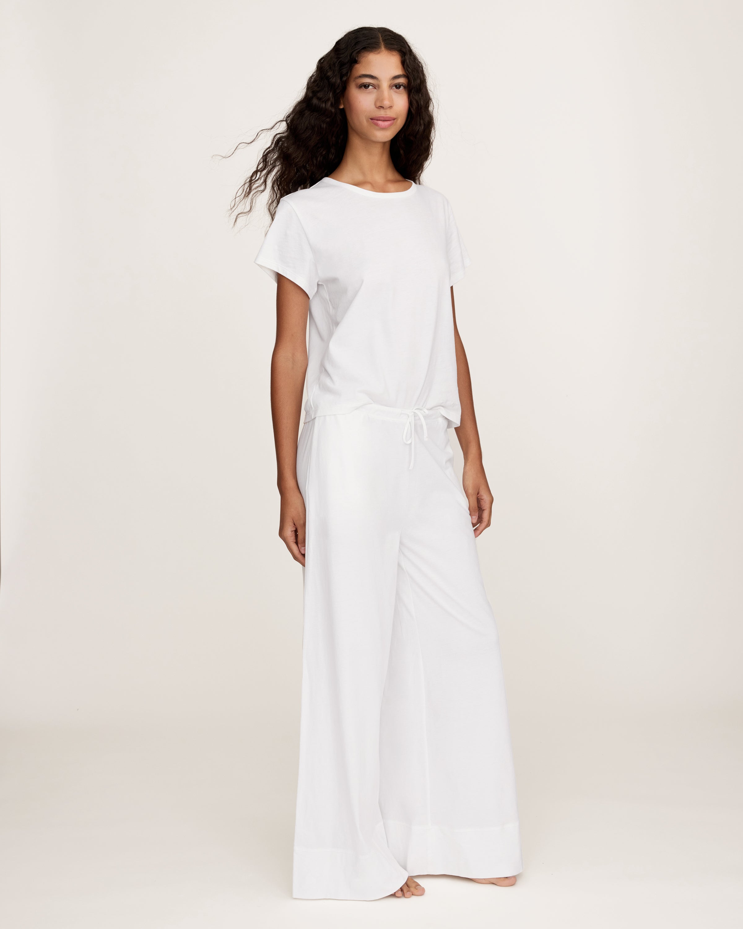 A person with long curly hair stands against a plain light background, wearing a loose-fitting white top and the Women's Pima Wide-Leg Lounge Pant in White, with one hand in a pocket.