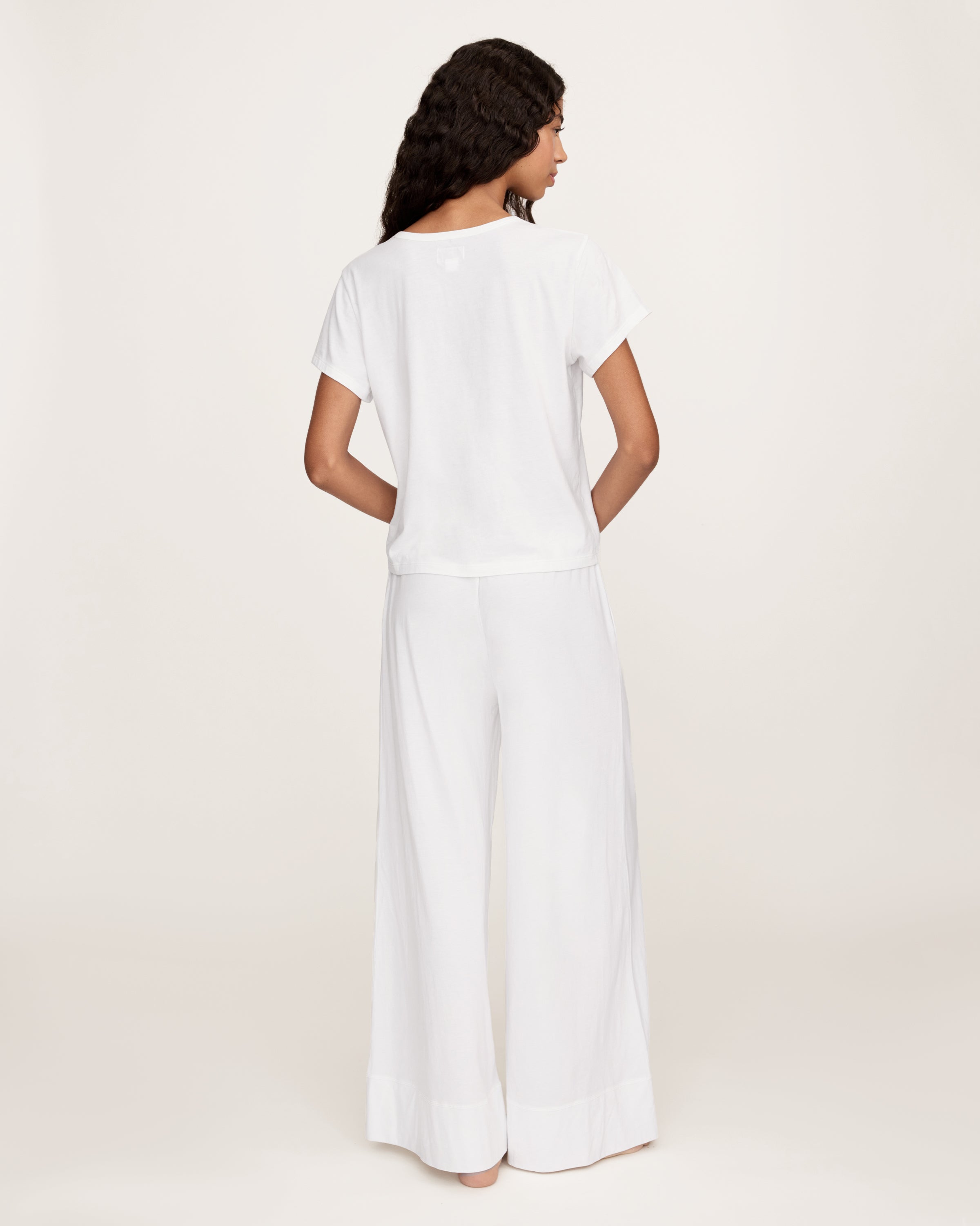 A person with long curly hair stands against a plain light background, wearing a loose-fitting white top and the Women's Pima Wide-Leg Lounge Pant in White, with one hand in a pocket.