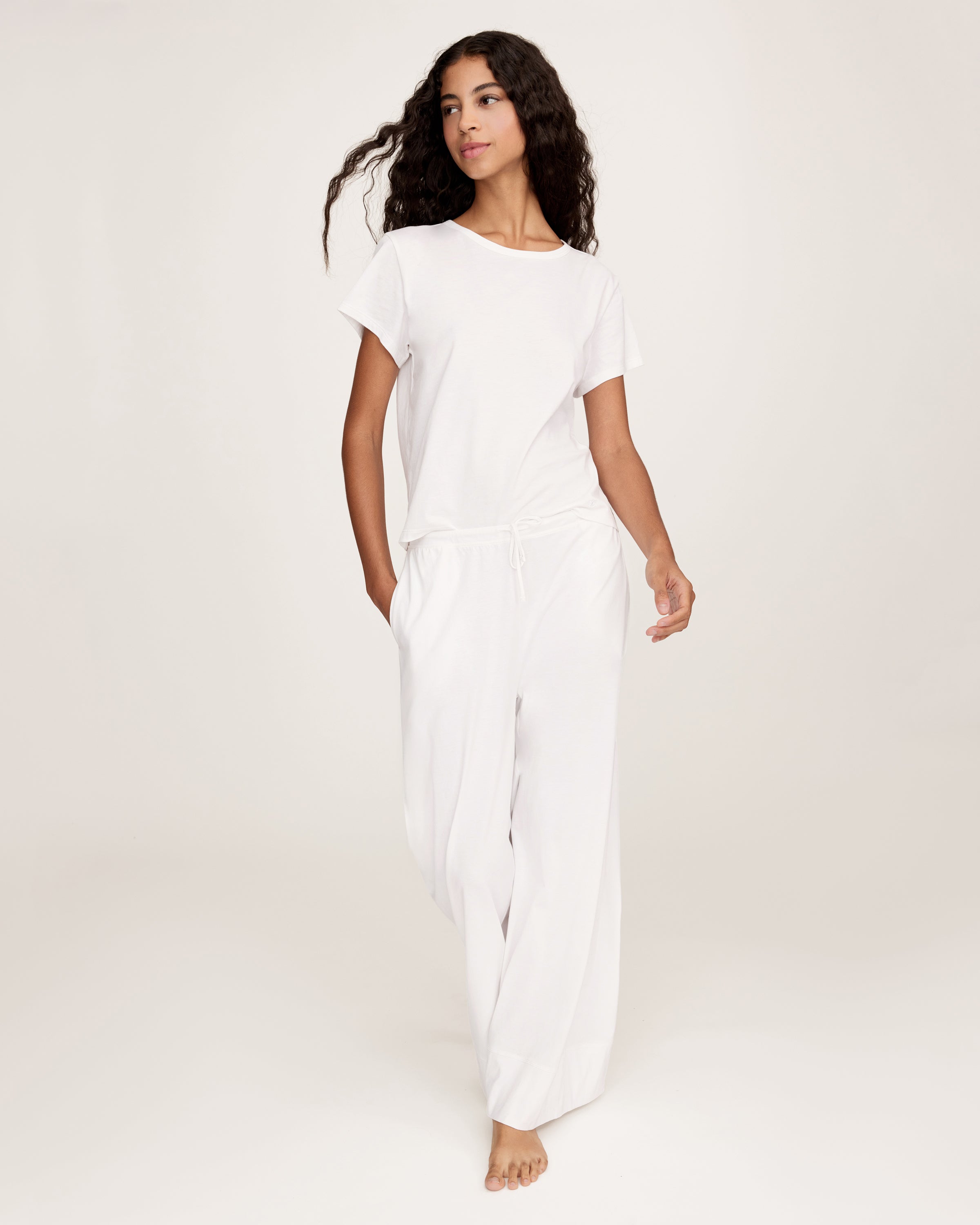 A person with long curly hair stands against a plain light background, wearing a loose-fitting white top and the Women's Pima Wide-Leg Lounge Pant in White, with one hand in a pocket.