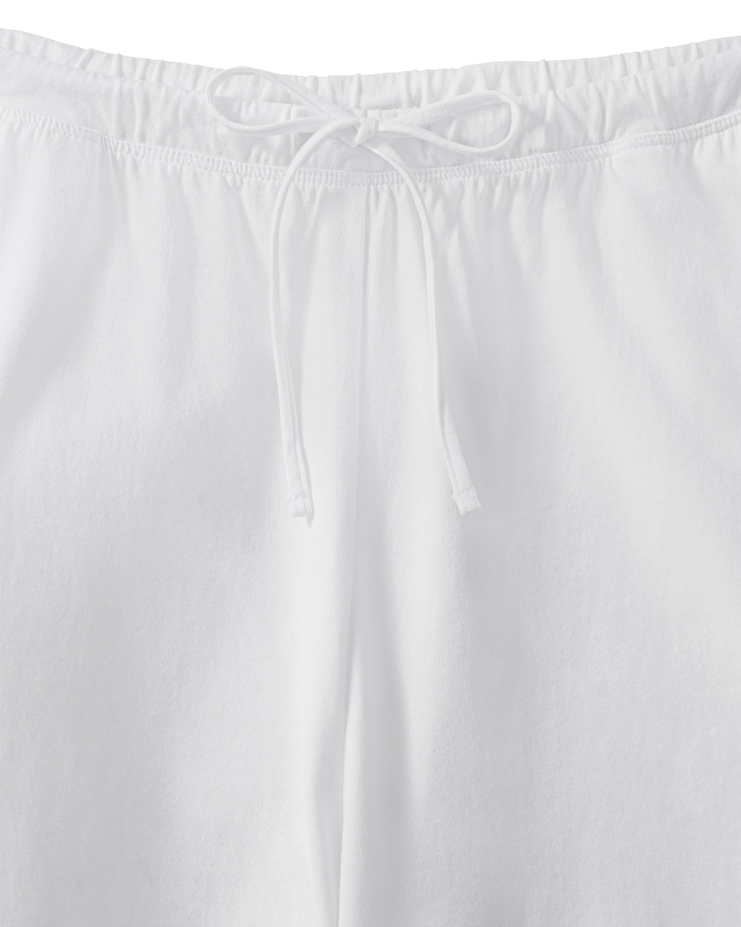 A person with long curly hair stands against a plain light background, wearing a loose-fitting white top and the Women's Pima Wide-Leg Lounge Pant in White, with one hand in a pocket.