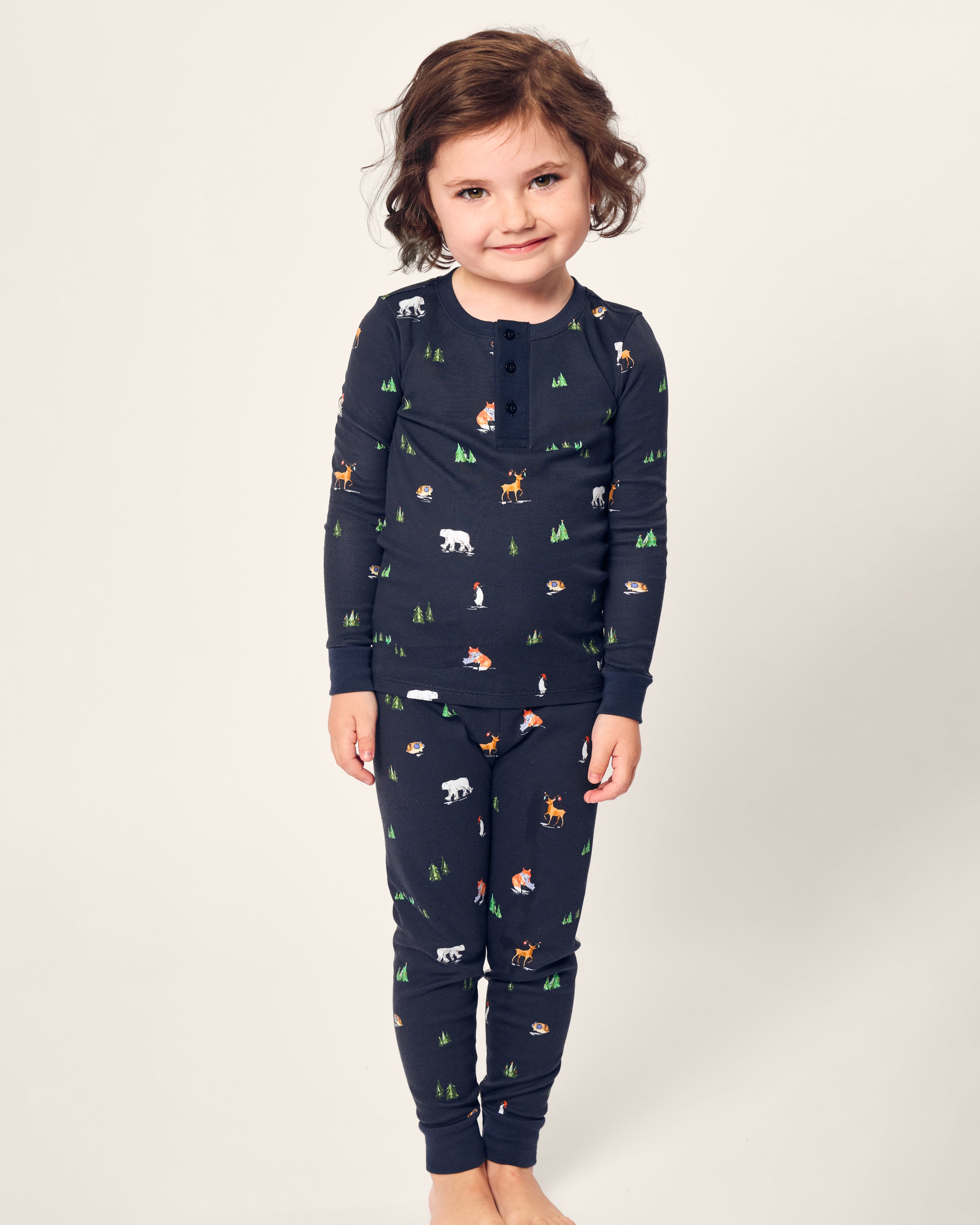 A young child smiles, clad in Petite Plumes cozy Kids Pima Snug Fit Pajama Set in Arctic Antics. The long sleeve navy blue top, adorned with small animal and forest prints, pops against a plain light background.