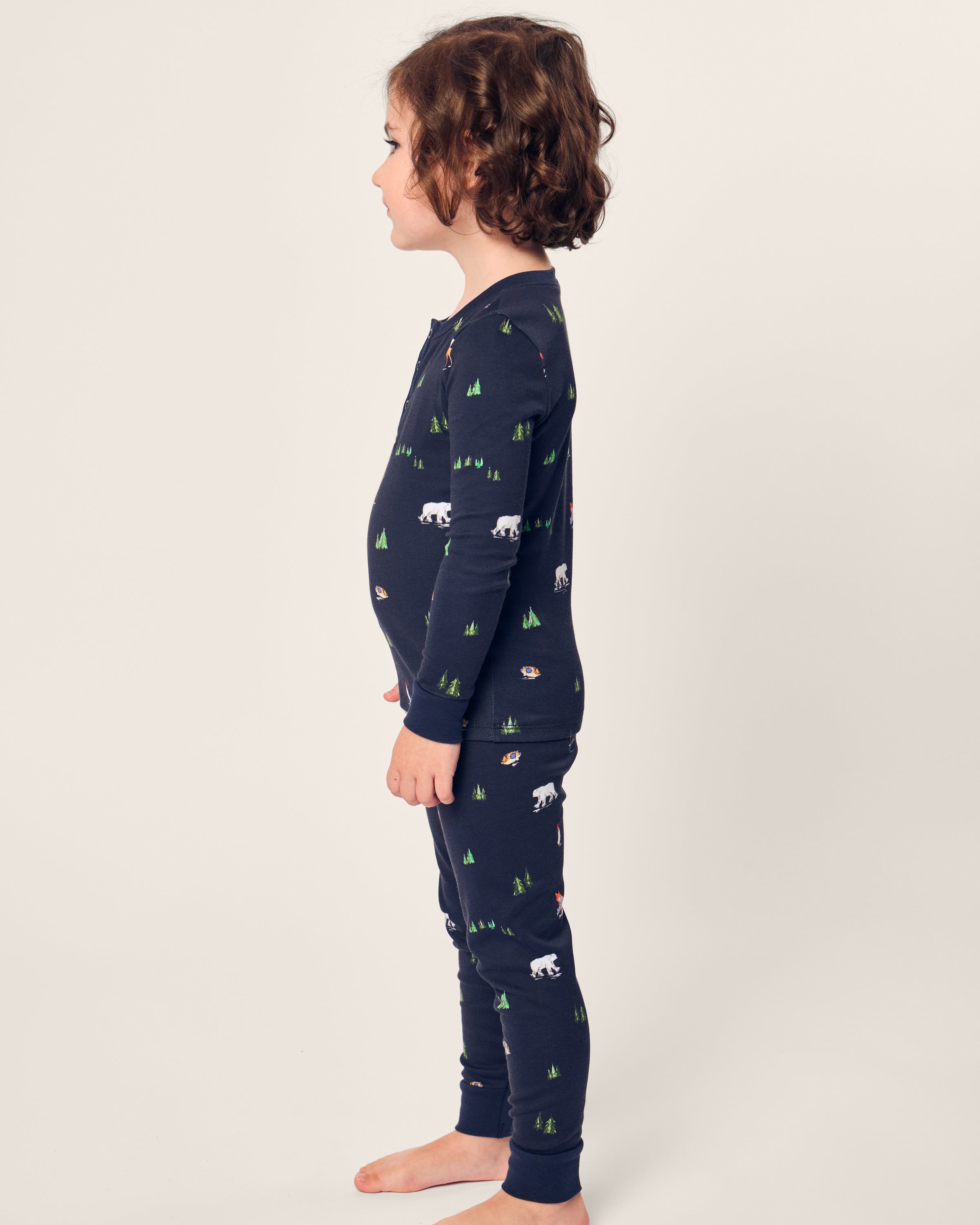 A child with curly hair stands sideways in Petite Plumes Kids Pima Snug Fit Pajama Set in Arctic Antics, featuring a navy nature-themed print of polar bears and trees on soft Pima cotton, with a cozy long sleeve top against a plain white background.