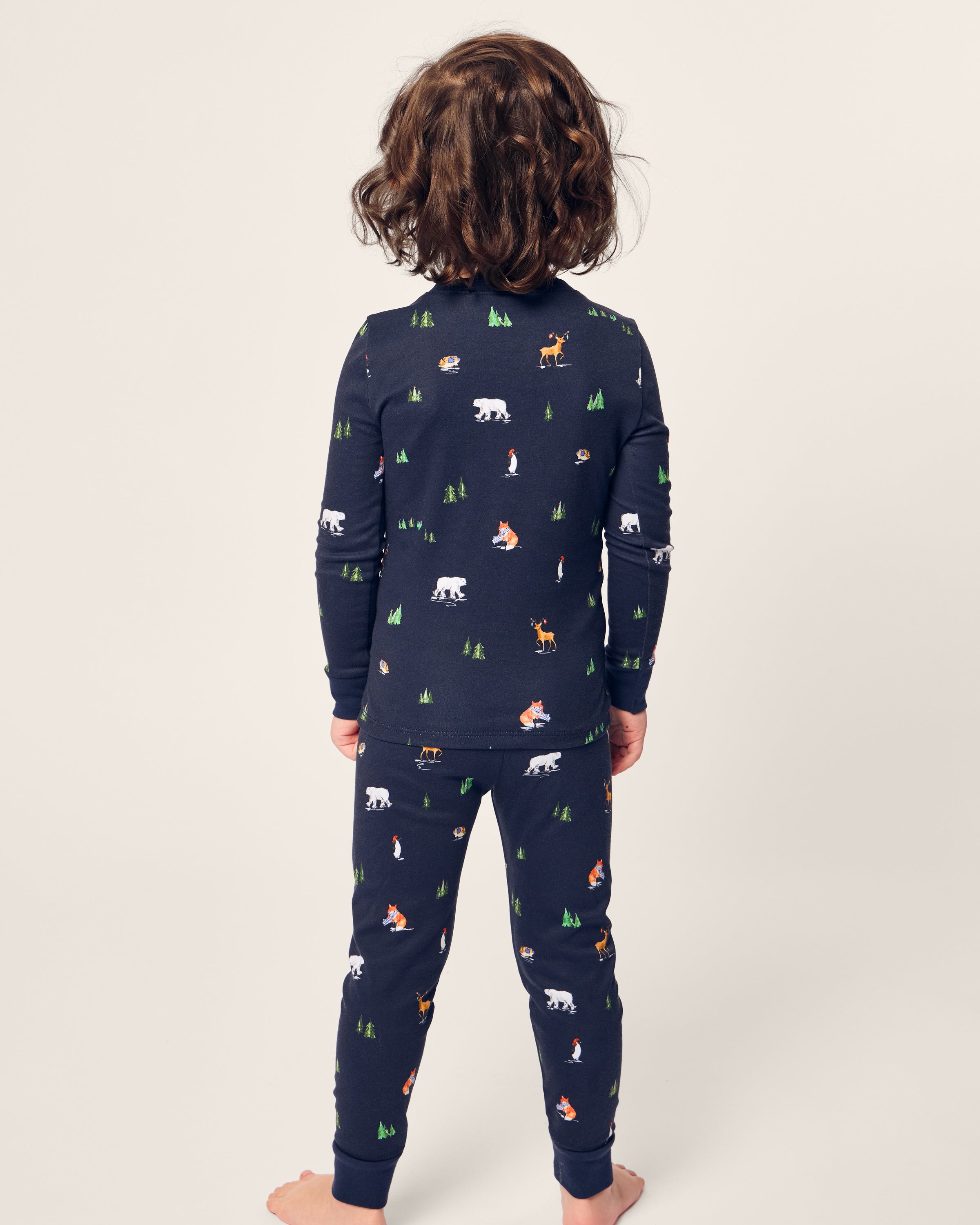 A child with curly hair stands barefoot, facing away, dressed in Petite Plumes Kids Pima Snug Fit Pajama Set in Arctic Antics. The long sleeve top and navy pajamas are made of soft Pima cotton, featuring charming bear and forest-themed motifs on a light background.