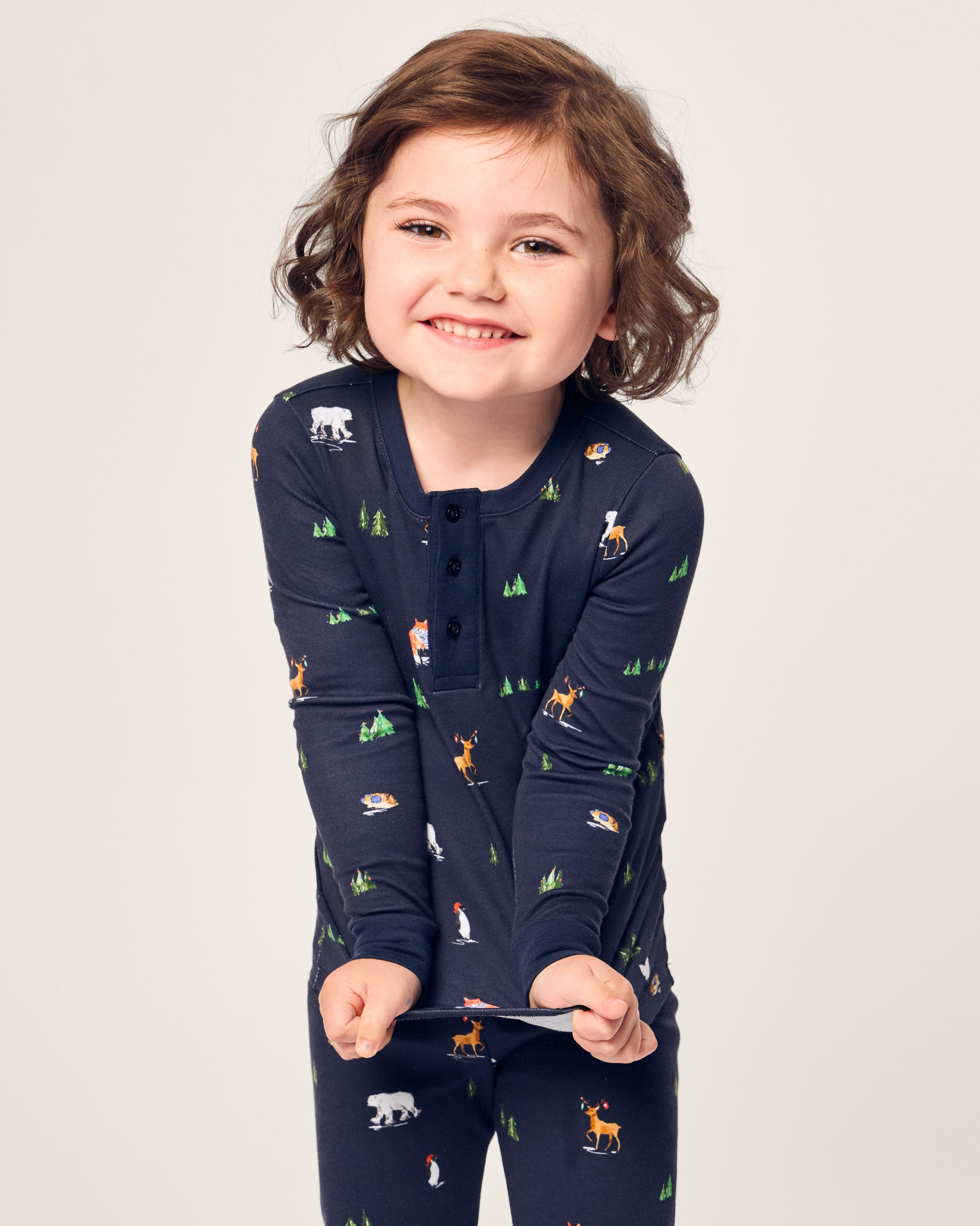 A child, smiling in bedtime fun, wears Petite Plumes Kids Pima Snug Fit Pajama Set in Arctic Antics. The dark pajamas feature bears, foxes, and penguins on a light background with trees. Theyre playfully stretching the soft Pima cotton long sleeve top.