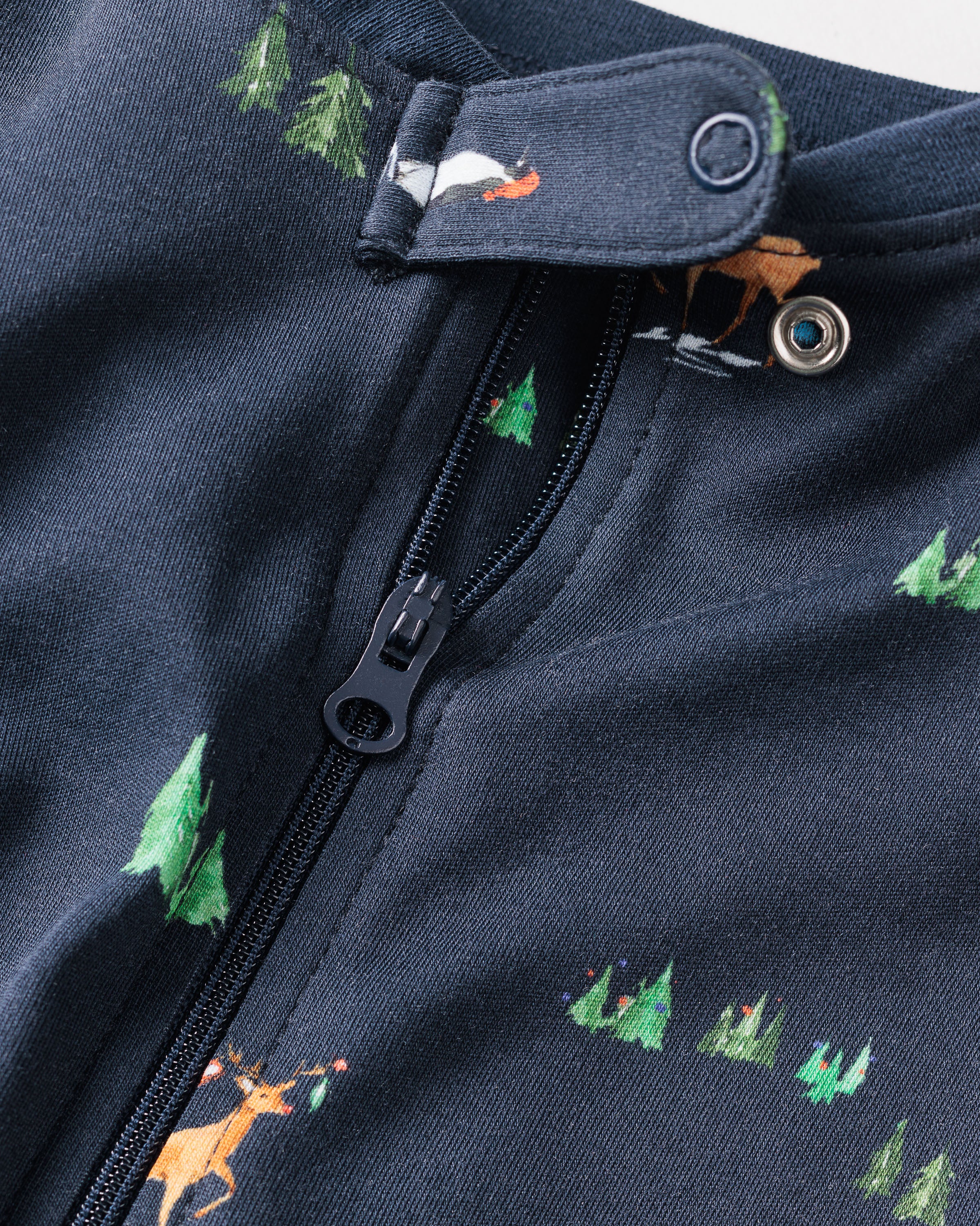 Close-up of the Kids Pima Snug Fit Pajama Set in Arctic Antics by Petite Plume, showcasing dark Pima cotton fabric with a zipper and button. It features festive tree and animal illustrations, including a red-nosed deer, embodying cozy holiday sleepwear.