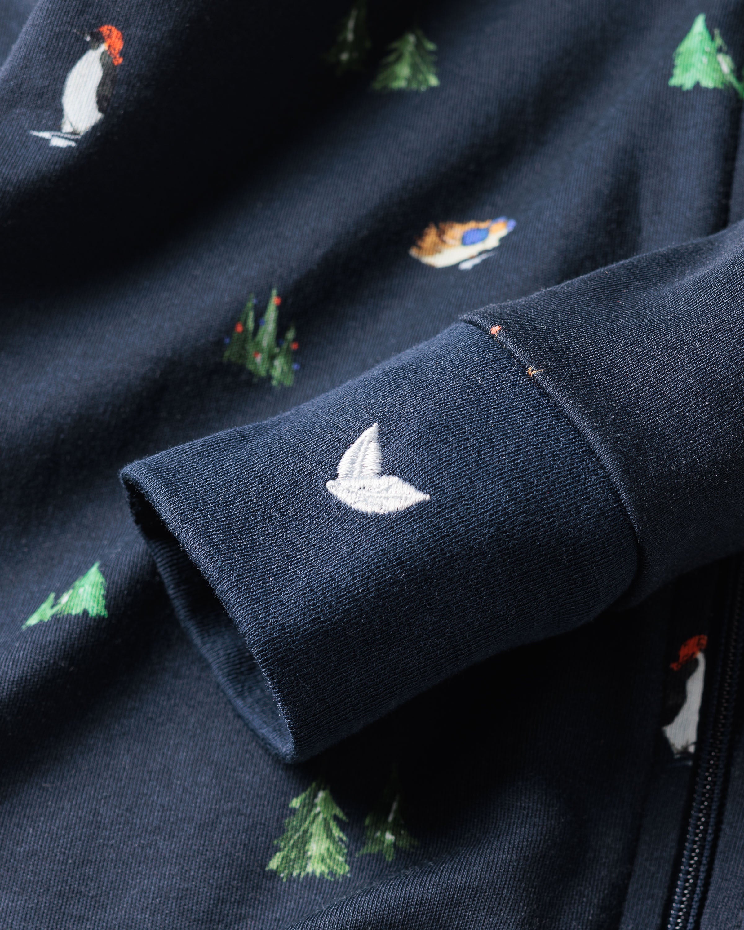 A close-up of the Petite Plume Kids Pima Snug Fit Pajama Set in Arctic Antics shows a navy fabric with detailed embroidery, including a white bird on the long sleeve top, penguins, and green Christmas trees. Perfect for cozy sleepwear fans who love intricate designs and luxurious comfort.