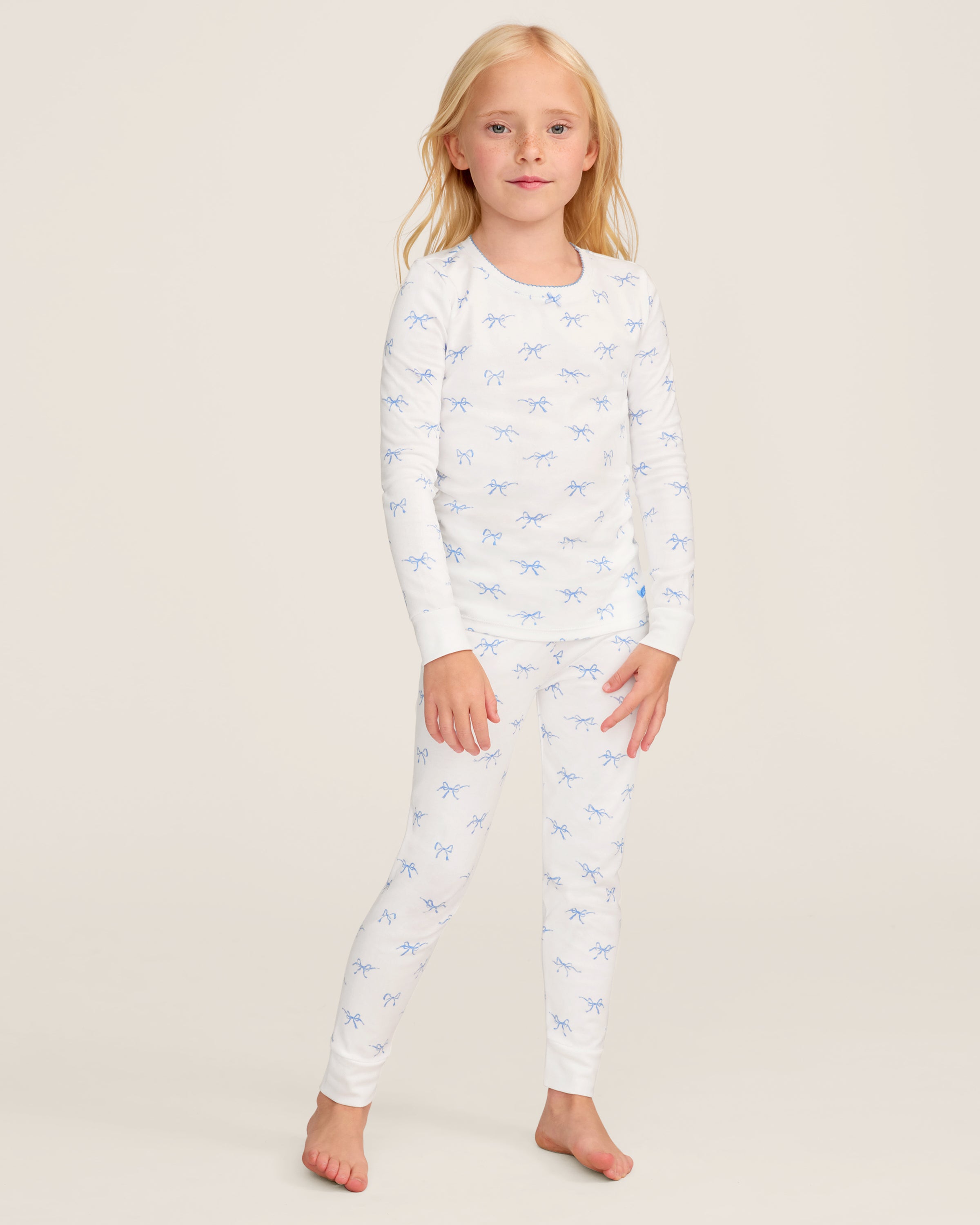 The Kid's Pima Snug Fit Pajama Set in Something Blue Bows features a long-sleeve white top and matching pants with a light blue bow pattern, crew neckline, and soft, comfortable fabric.