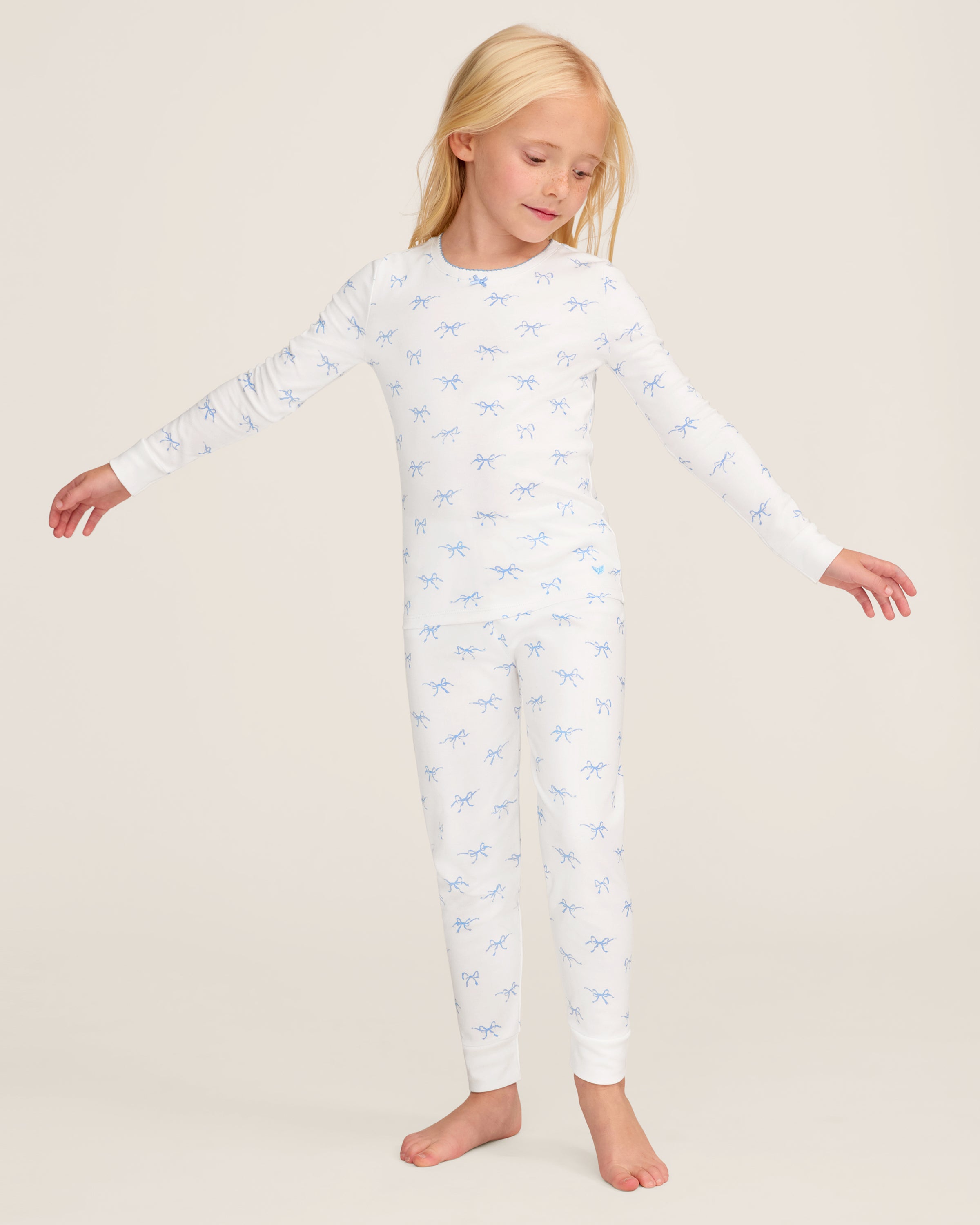 The Kid's Pima Snug Fit Pajama Set in Something Blue Bows features a long-sleeve white top and matching pants with a light blue bow pattern, crew neckline, and soft, comfortable fabric.