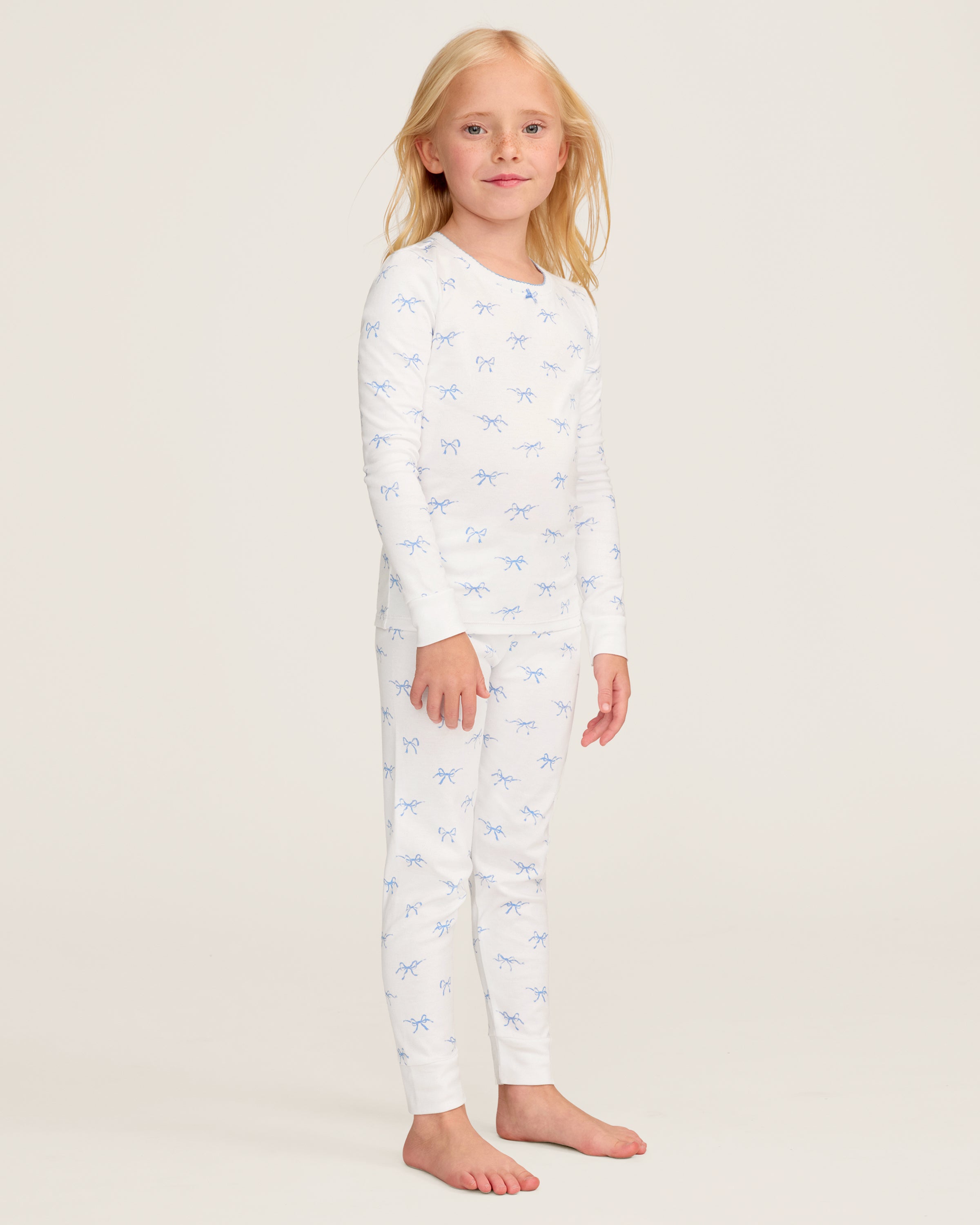 The Kid's Pima Snug Fit Pajama Set in Something Blue Bows features a long-sleeve white top and matching pants with a light blue bow pattern, crew neckline, and soft, comfortable fabric.