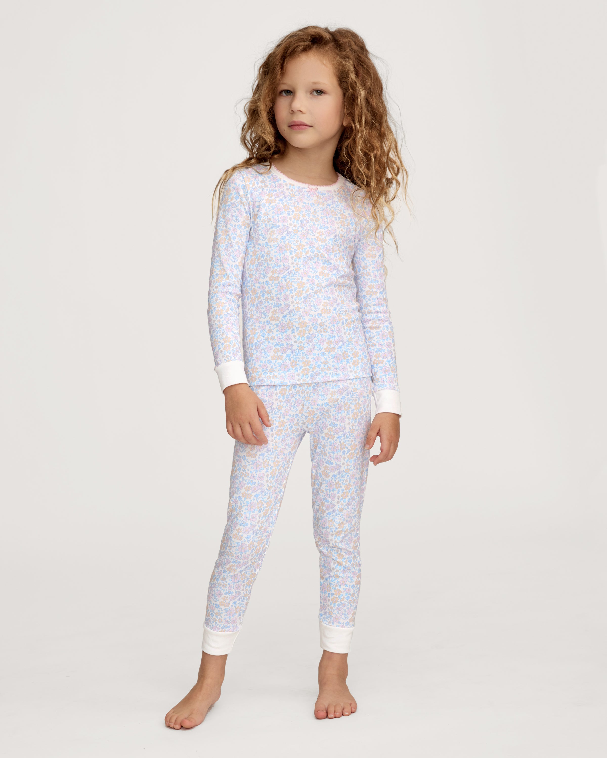 A young girl with long, wavy hair stands barefoot on a plain background, wearing the Kid's Pima Snug Fit Pajama Set in Fleur Blanche. She looks slightly to the side with a relaxed expression.