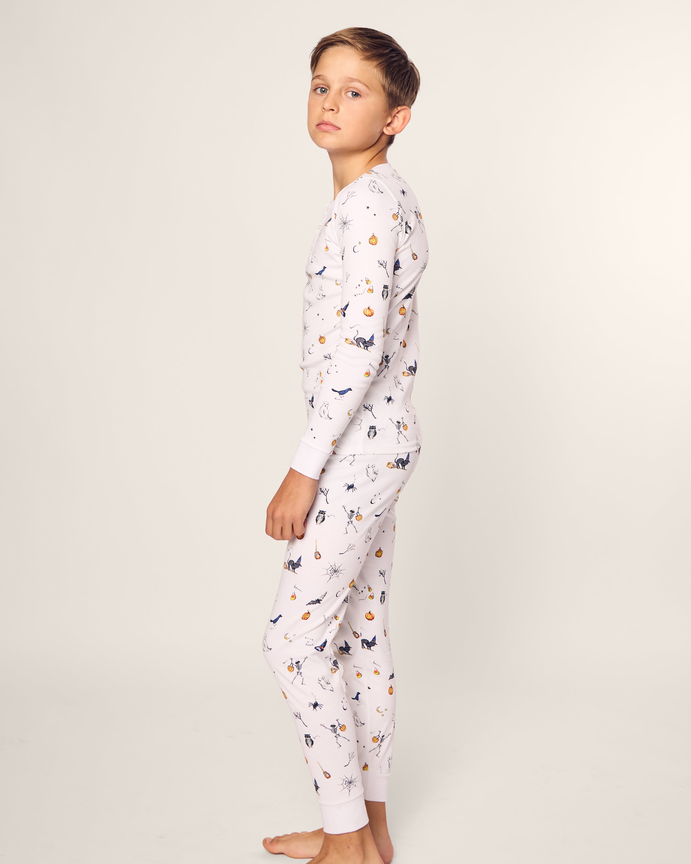 A young boy stands sideways, looking at the camera in his luxurious Petite Plume Kids Pima Snug Fit Pajama Set in Trick or Treat, adorned with playful animal and musical note prints, against a plain off-white background.