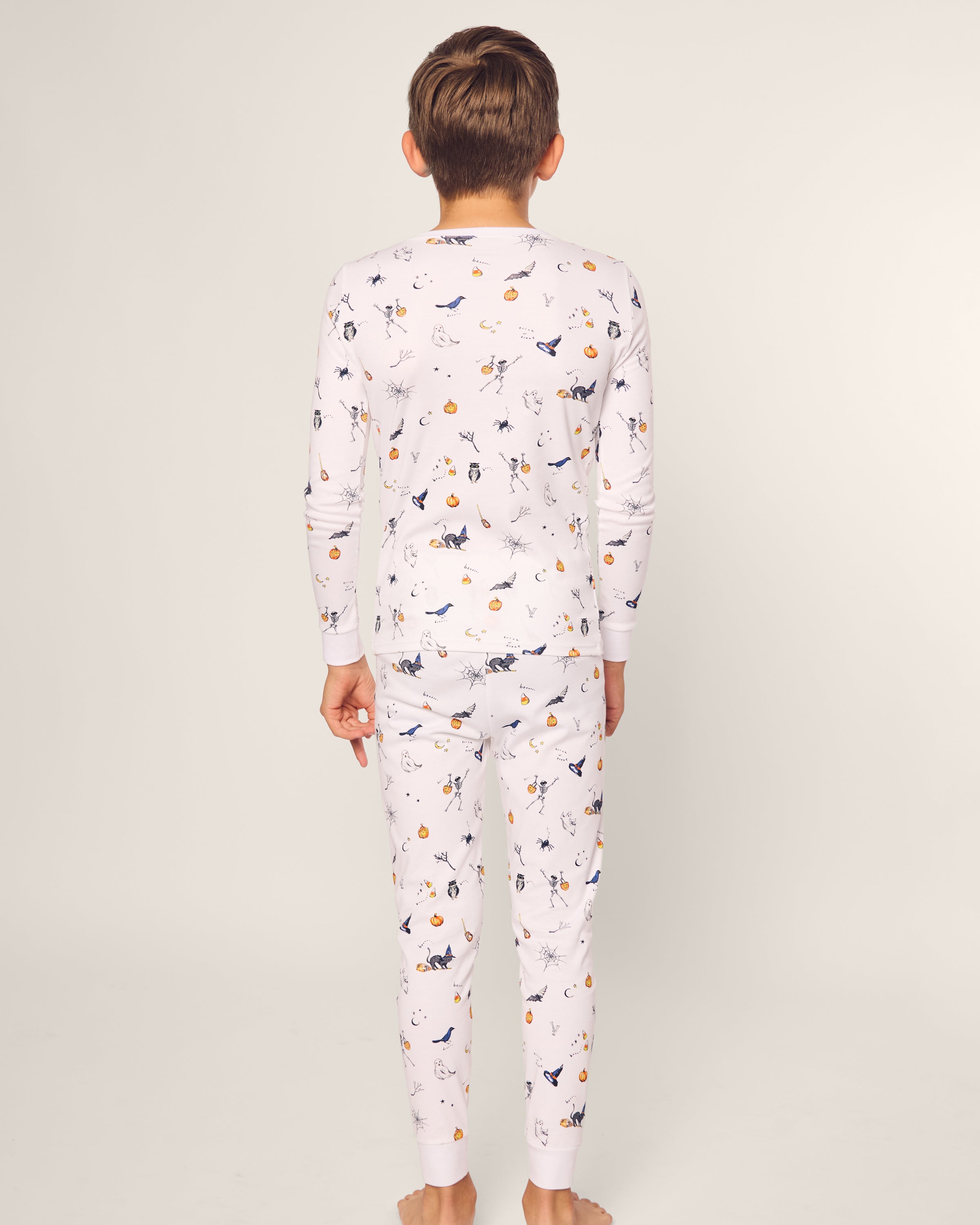 A child stands barefoot and facing away, dressed in the elegant Kids Pima Snug Fit Pajama Set in Trick or Treat by Petite Plume. The set features a long sleeve top and pants with colorful illustrations on white Pima cotton, against a plain off-white background showcasing comfort and style.