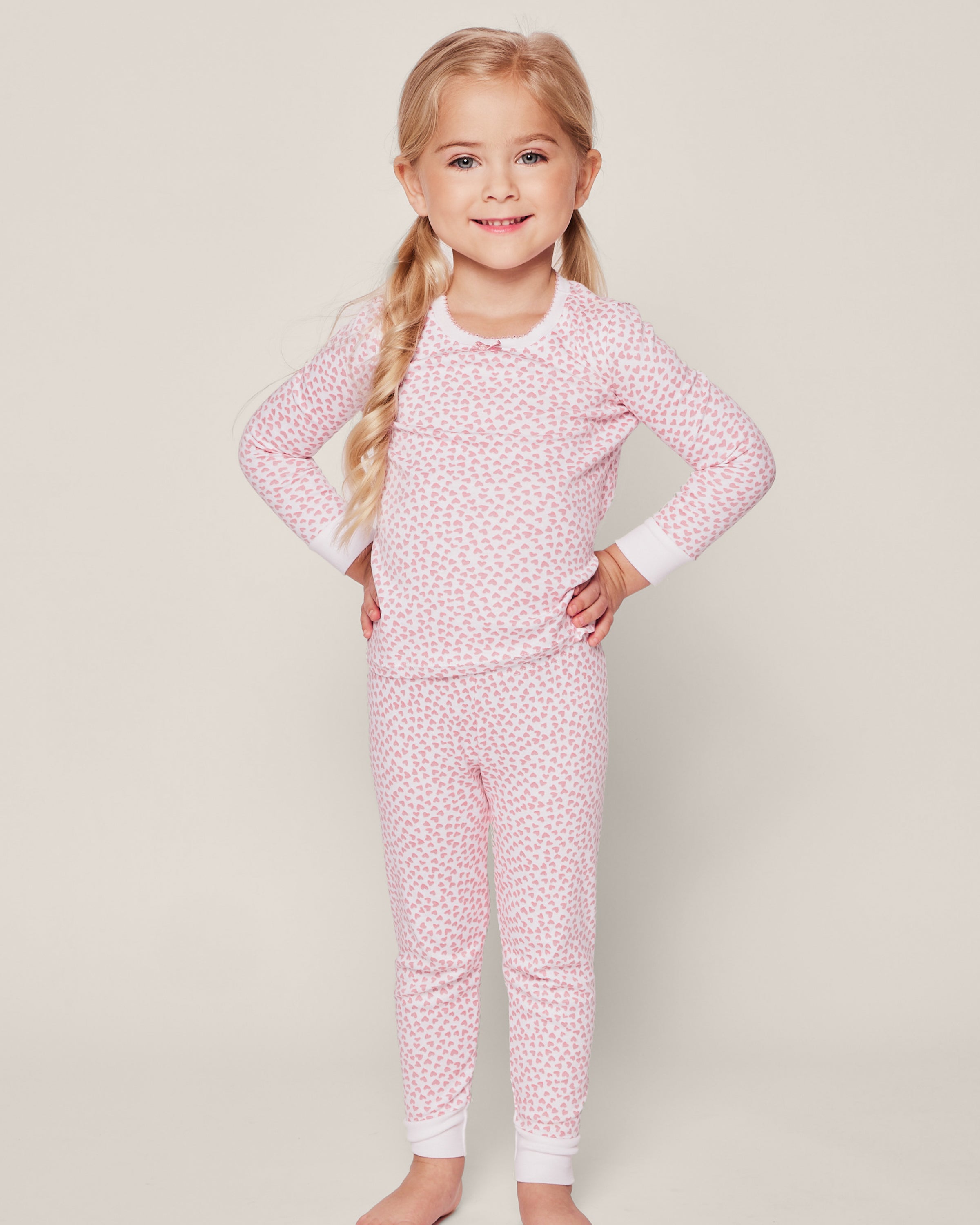 A young girl with long blonde hair smiles, wearing Petite Plumes Kids Pima Snug Fit Pajama Set in Sweethearts. The matching pink pajamas feature a white floral pattern and are made from soft Pima cotton, complementing her cheerful demeanor against the plain white background.