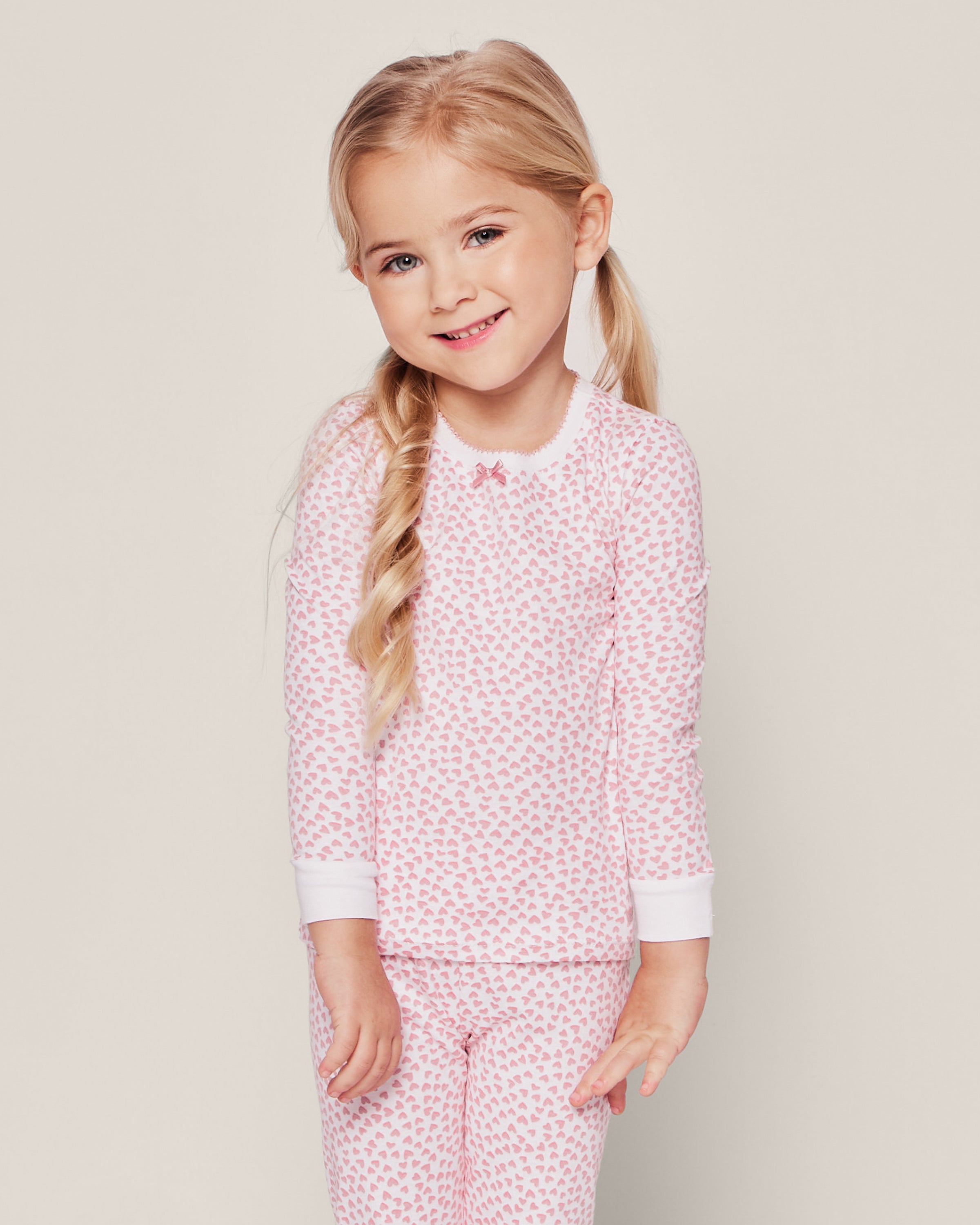 A young girl with long blonde hair in a side braid smiles in Petite Plumes luxurious Kids Pima Snug Fit Pajama Set in Sweethearts, featuring pink pajamas with small heart patterns, including a cozy long-sleeve top and pants, as she stands against a plain background.