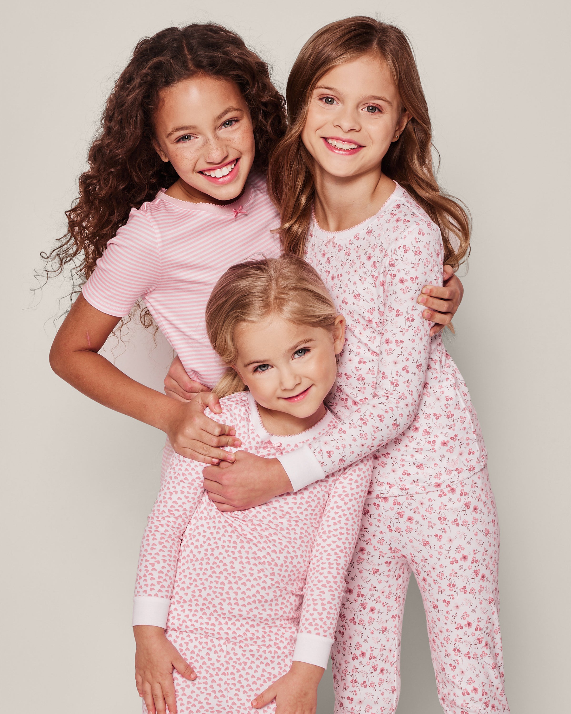 Three smiling children embrace each other against a plain light background, wearing Petite Plumes Kids Pima Snug Fit Pajama Set in Sweethearts—a luxurious pink and white sleepwear set with a long sleeve top and pants, perfect for comfort.