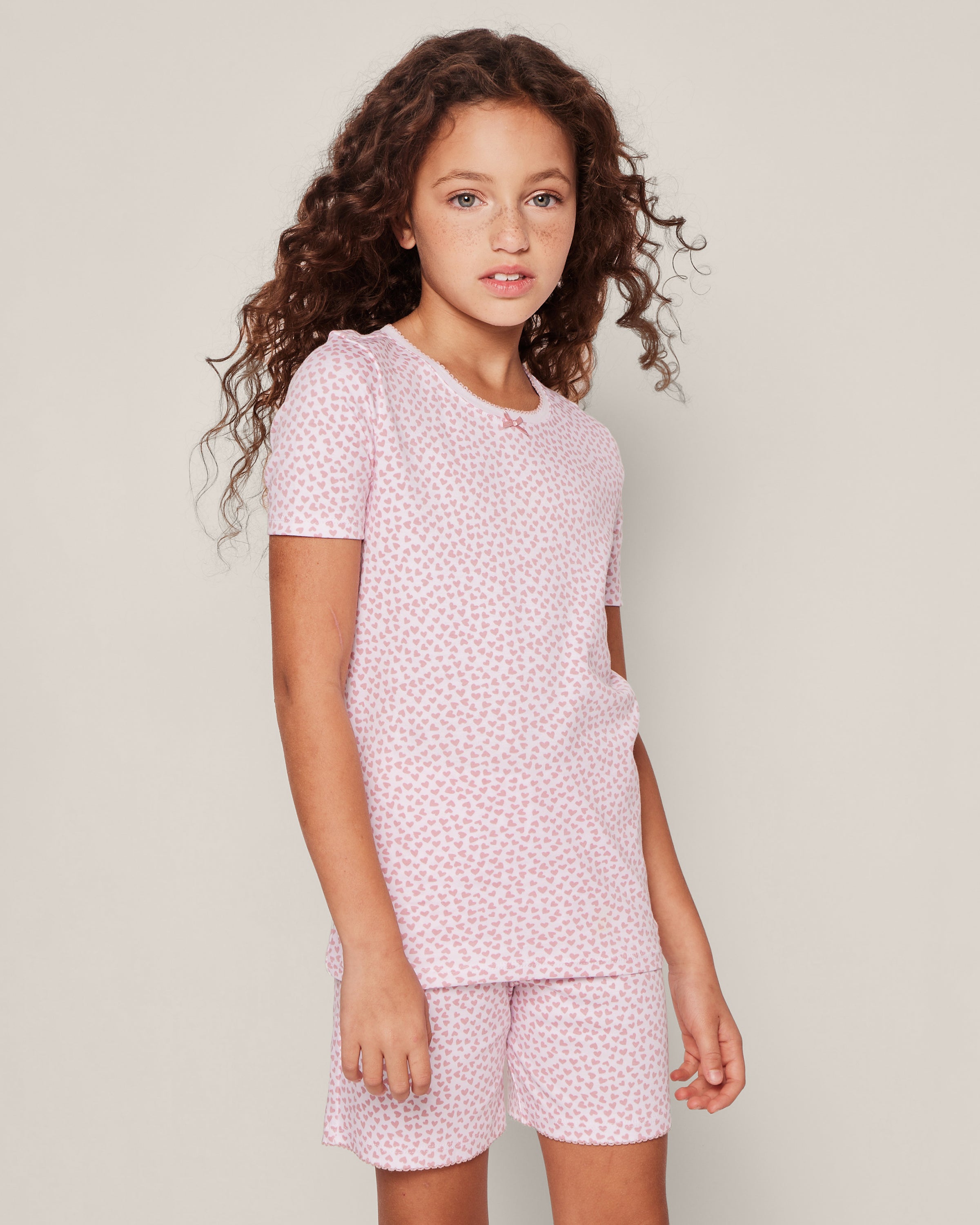 A young girl with long, curly hair wears Petite Plumes Kids Pima Snug Fit Pajama Short Set in Sweethearts. She stands against a plain background, looking forward with a neutral expression.