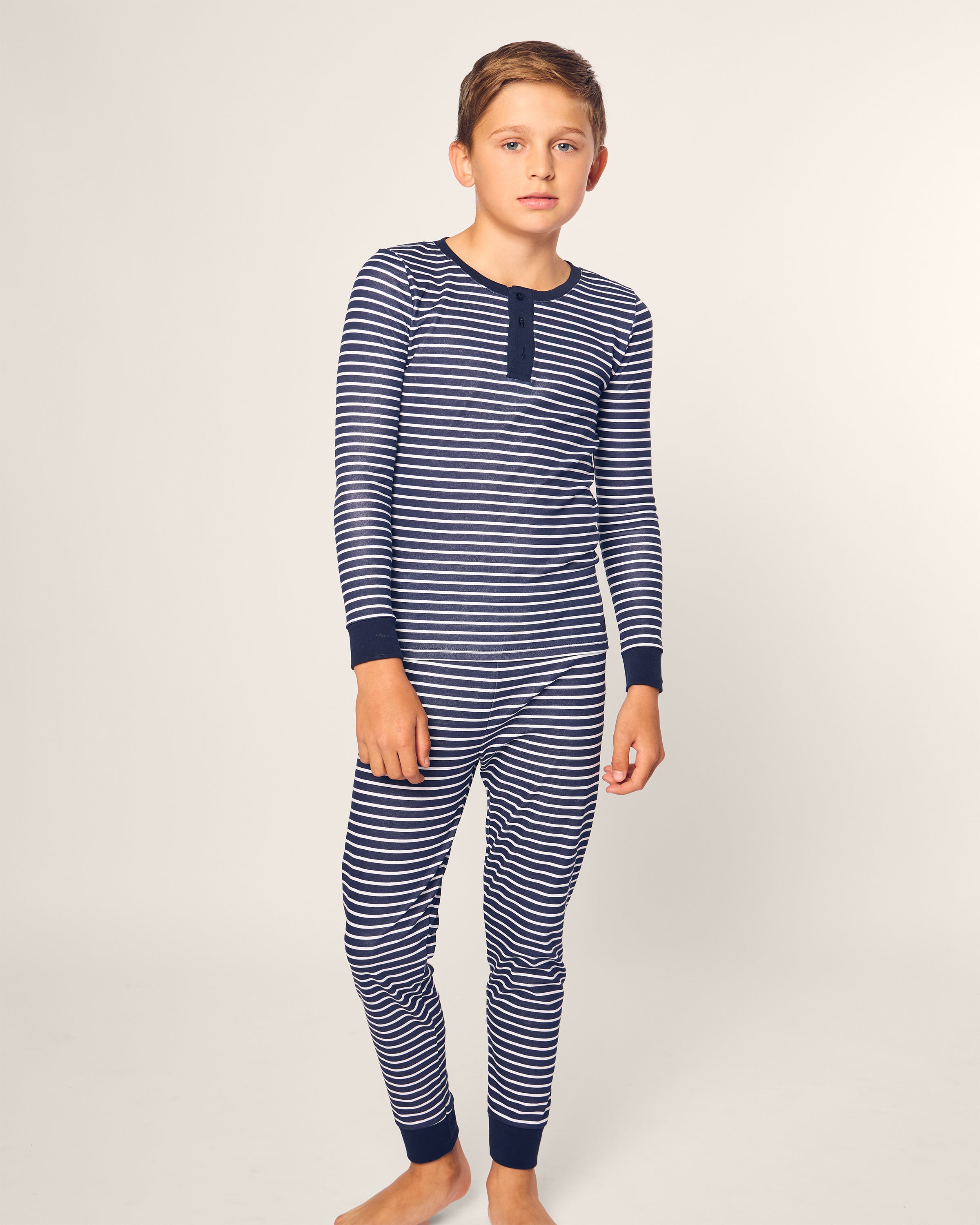 A young boy stands barefoot against a plain background, wearing Petite Plumes Kids Pima Snug Fit Pajama Set in Navy Stripe with long sleeves, dark cuffs, and a short buttoned placket. His expression is neutral as he looks toward the camera.