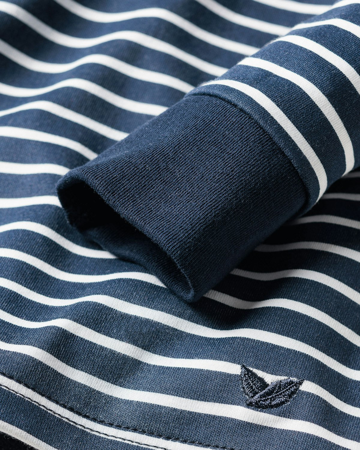 The Petite Plume Kids Pima Snug Fit Pajama Set in Navy Stripe shows a close-up of its fabric featuring white stripes, a sleeve cuff, and an embroidered boat near the hem. Made from luxurious Pima Cotton, this set ensures comfort and style for restful nights.