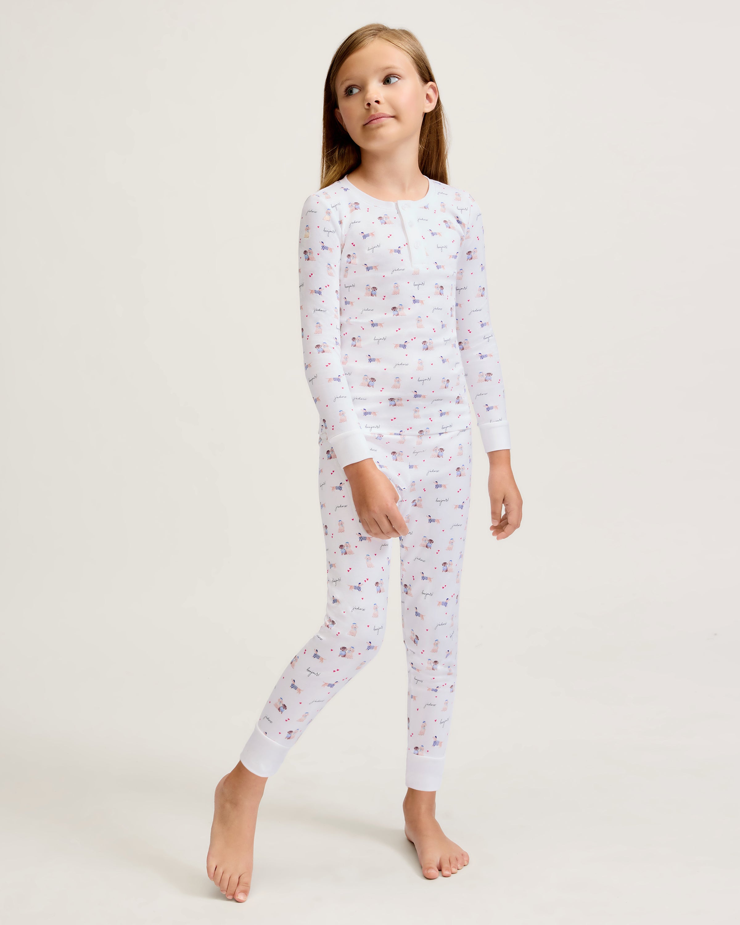 The Kid's Pima Snug Fit Pajama Set in Le Woof is a white long-sleeve pajama set with matching pants, featuring a colorful all-over cartoon dog and bone print, plus cuffed sleeves and ankles for a snug fit.