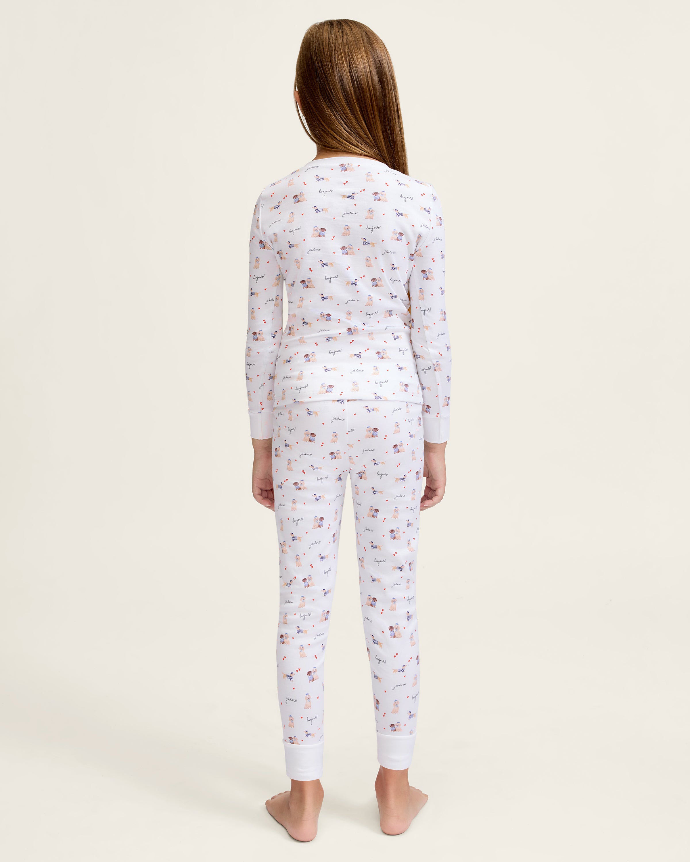 The Kid's Pima Snug Fit Pajama Set in Le Woof is a white long-sleeve pajama set with matching pants, featuring a colorful all-over cartoon dog and bone print, plus cuffed sleeves and ankles for a snug fit.