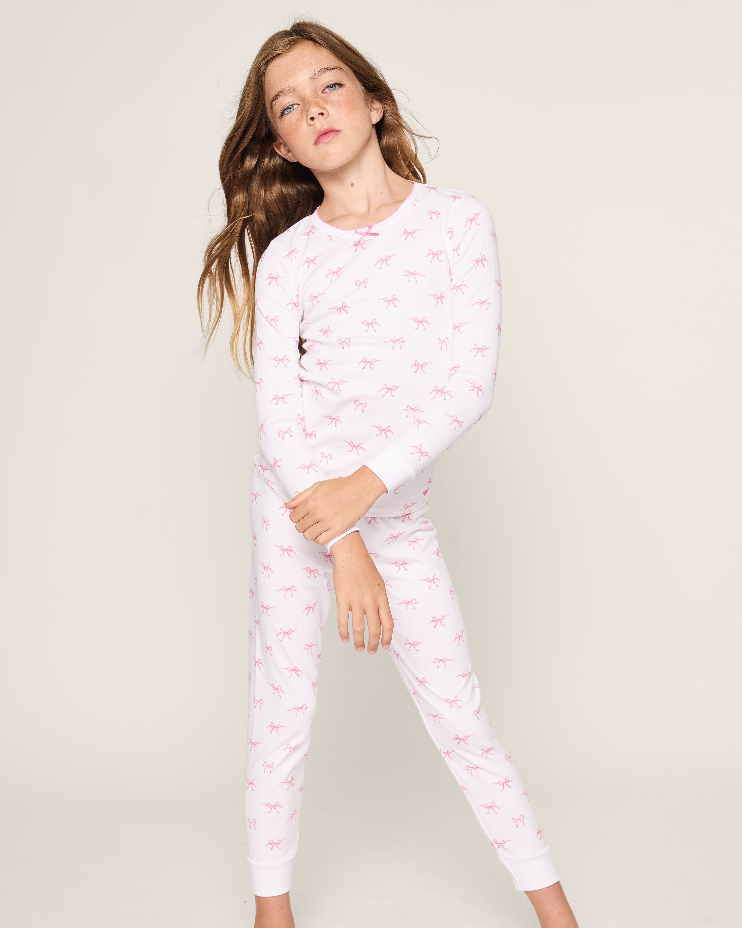 A confident young girl with long brown hair wears the Petite Plume Kids Pima Snug Fit Pajama Set in Blushing Bows, featuring a long sleeve top and pants made from soft Pima cotton with small pink patterns against a plain light-colored background.