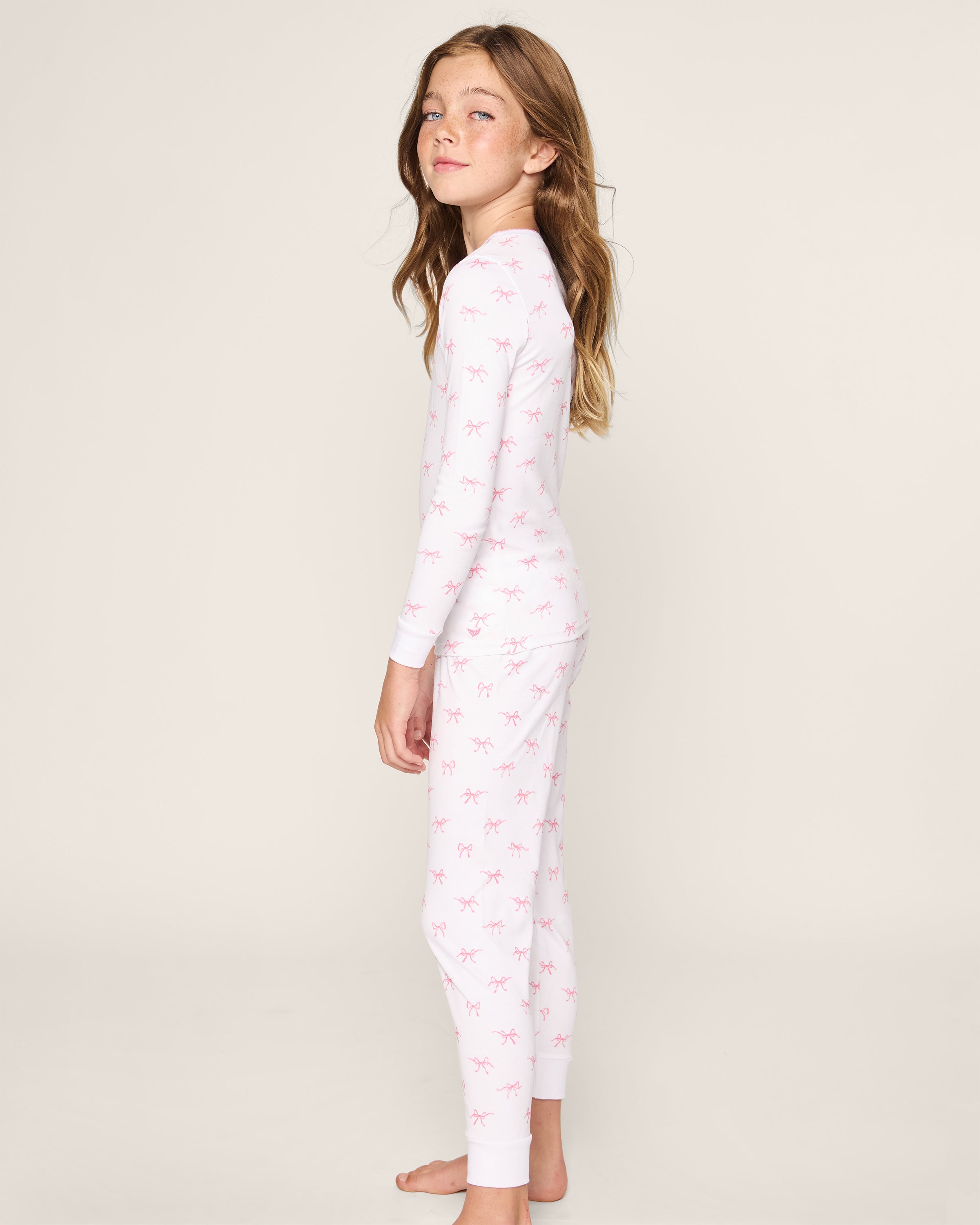 A young girl with long hair wears the Kids Pima Snug Fit Pajama Set in Blushing Bows by Petite Plume, posing against a plain background, exuding cozy elegance.