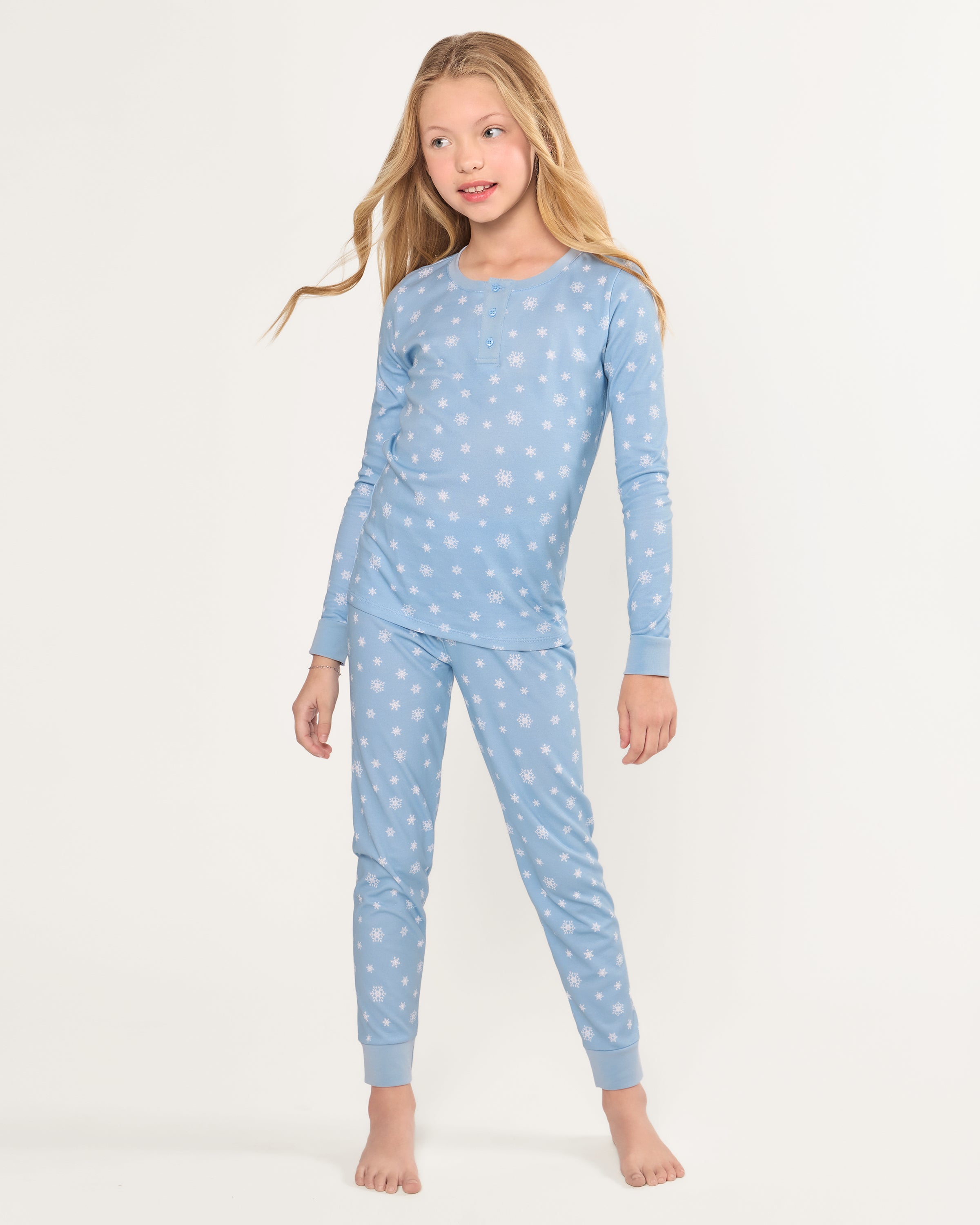 The Kid's Pima Snug Fit Pajama Set in Periwinkle Wonderland features light blue, long-sleeve pajamas with white snowflakes, buttoned neckline, matching elastic waist pants, and cuffed ankles. Shown laid flat on a white background.
