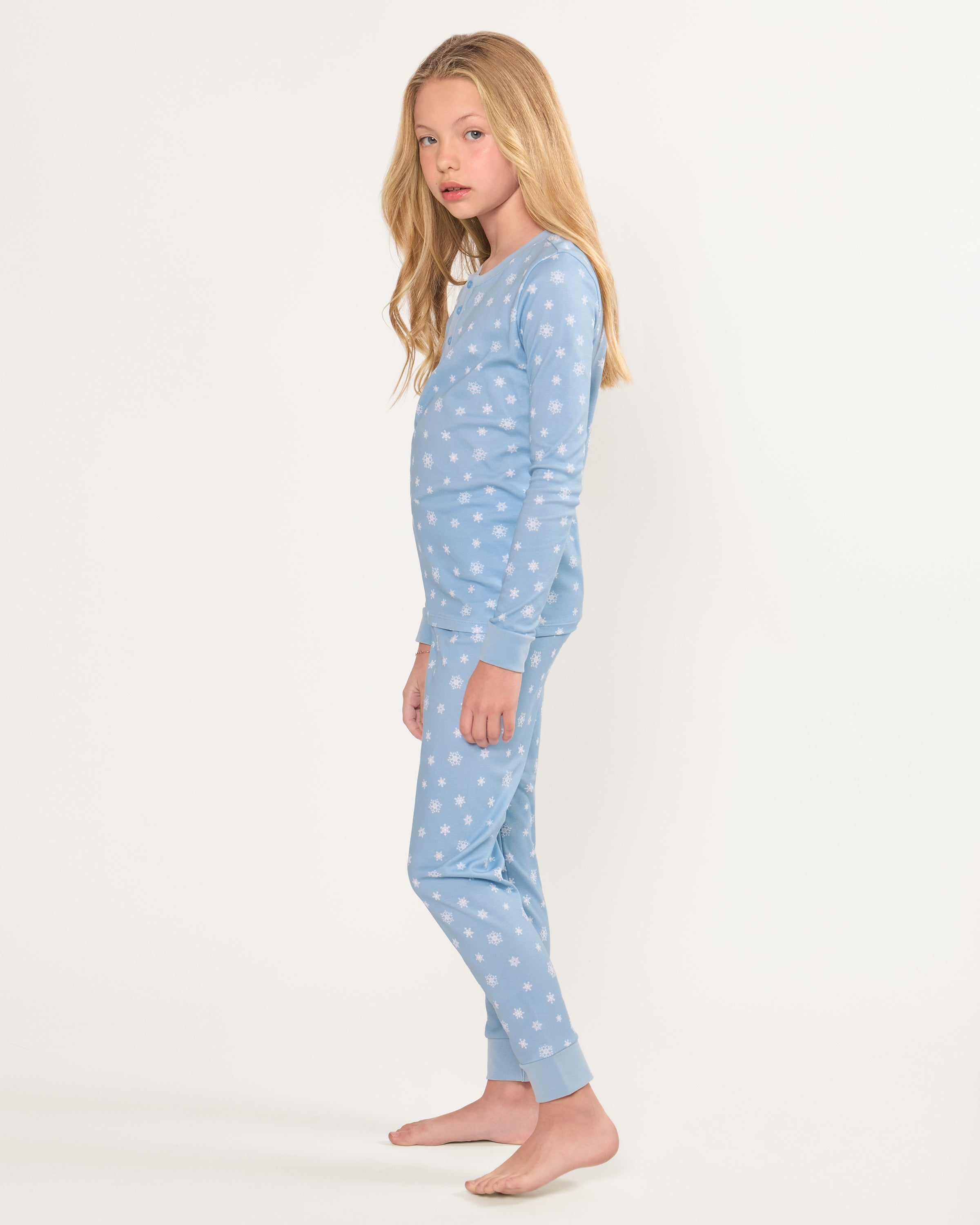 The Kid's Pima Snug Fit Pajama Set in Periwinkle Wonderland features light blue, long-sleeve pajamas with white snowflakes, buttoned neckline, matching elastic waist pants, and cuffed ankles. Shown laid flat on a white background.