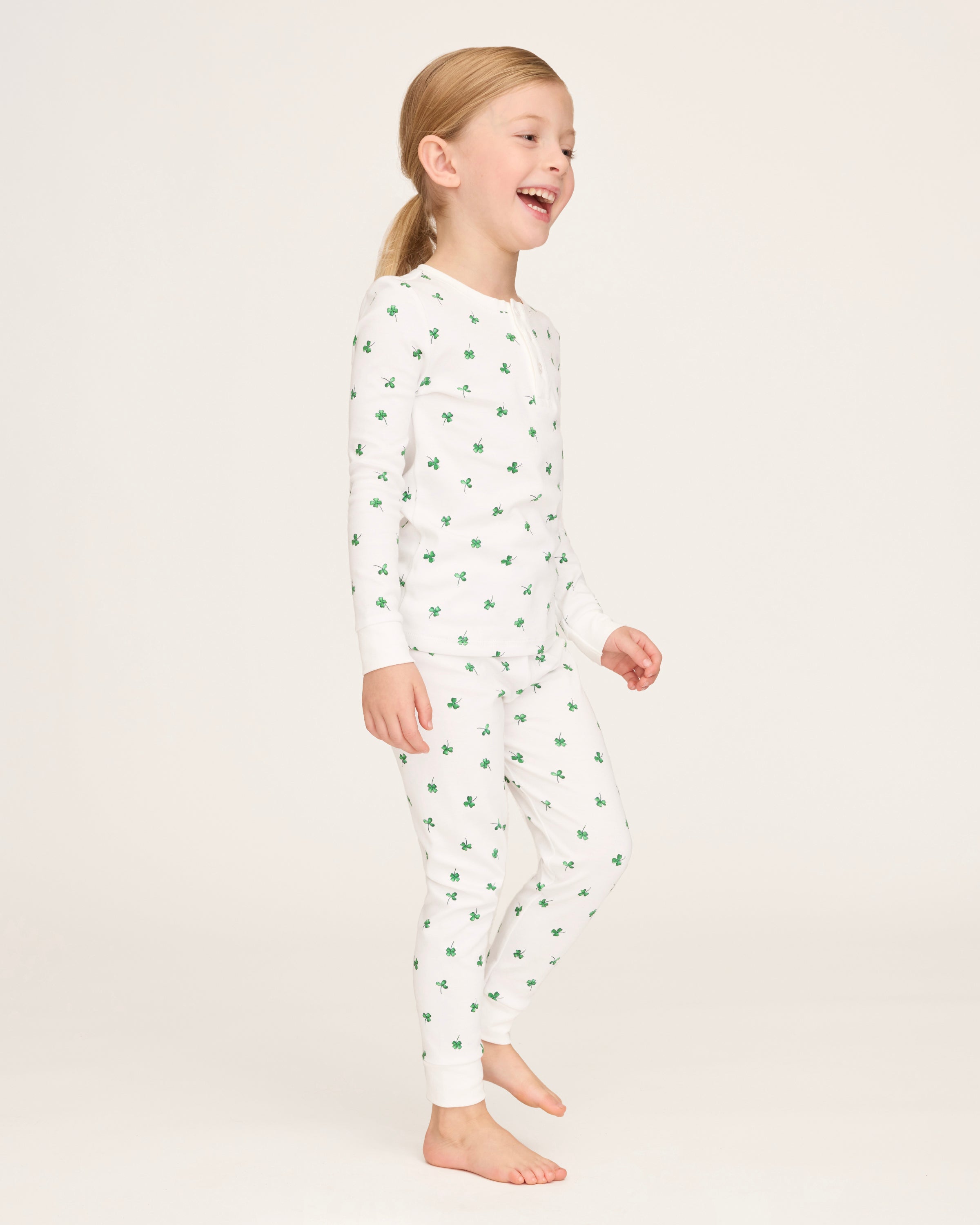 A young child with blonde hair smiles barefoot against a plain light background, dressed in the Kid's Pima Snug Fit Pajama Set in Shamrocks—white long-sleeve pajamas featuring small green shamrock patterns.