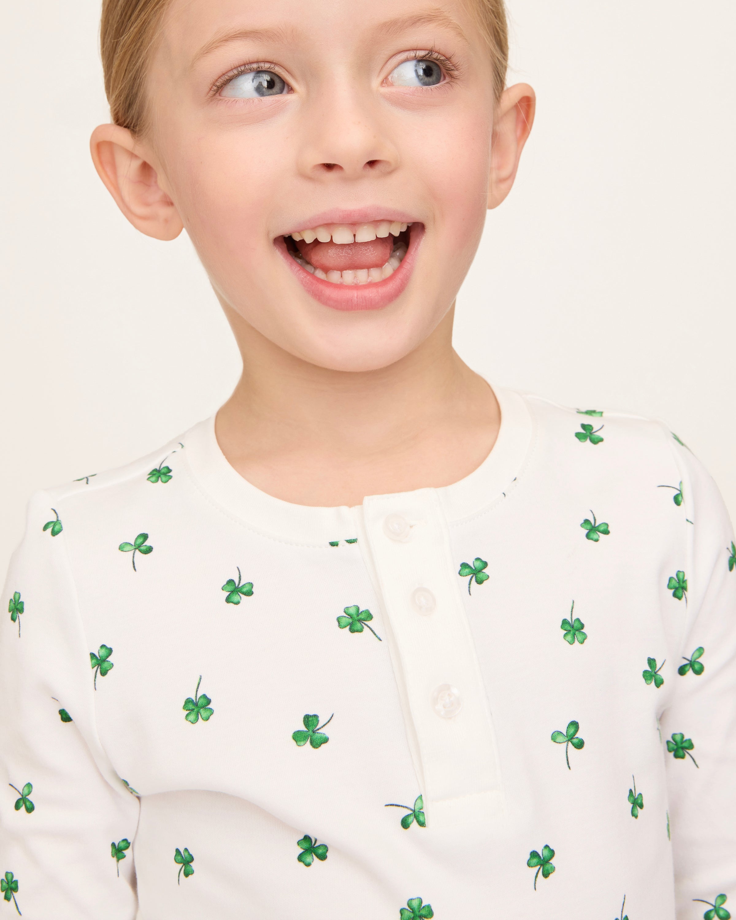 A young child with blonde hair smiles barefoot against a plain light background, dressed in the Kid's Pima Snug Fit Pajama Set in Shamrocks—white long-sleeve pajamas featuring small green shamrock patterns.