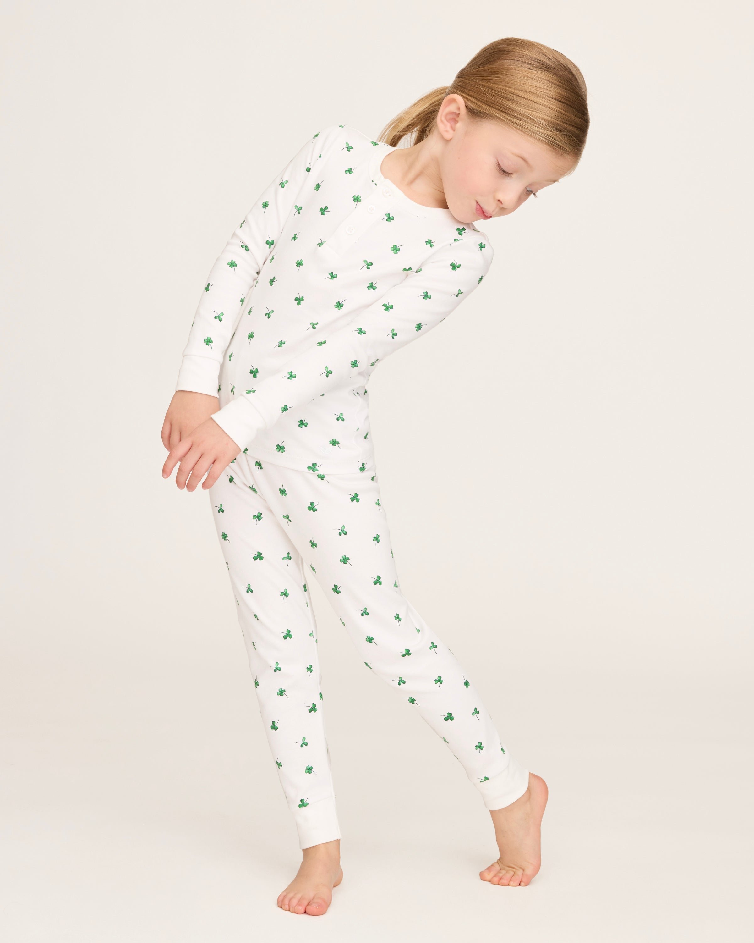 A young child with blonde hair smiles barefoot against a plain light background, dressed in the Kid's Pima Snug Fit Pajama Set in Shamrocks—white long-sleeve pajamas featuring small green shamrock patterns.