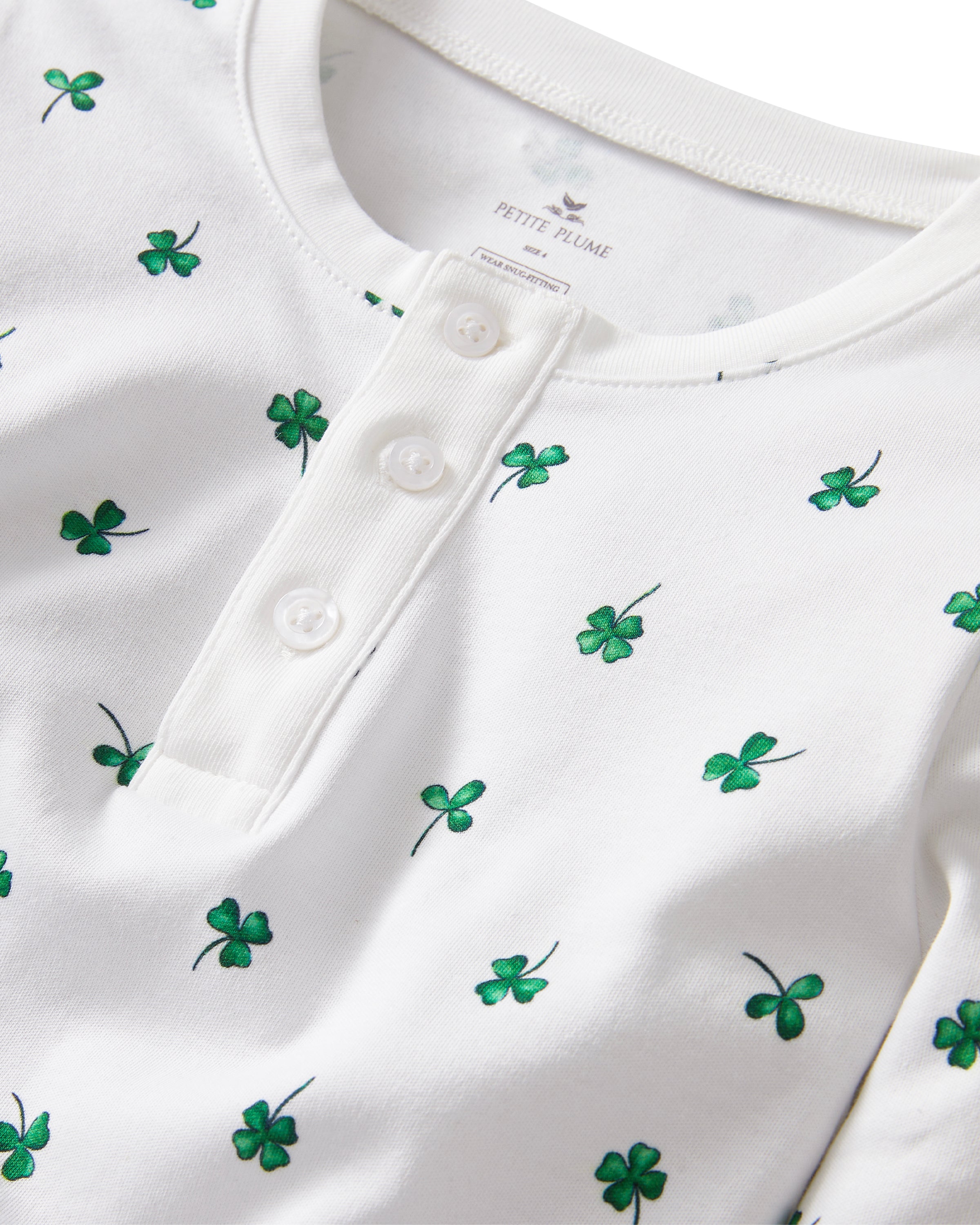 A young child with blonde hair smiles barefoot against a plain light background, dressed in the Kid's Pima Snug Fit Pajama Set in Shamrocks—white long-sleeve pajamas featuring small green shamrock patterns.