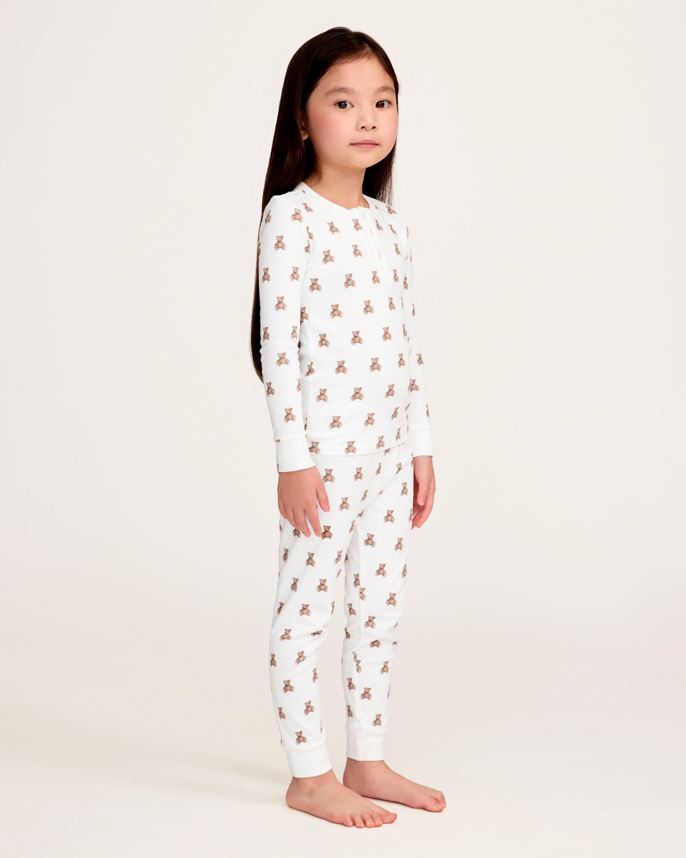 The Kid's Pima Snug Fit Pajama Set in Teddy Tales features a white long-sleeve buttoned top and elastic waist pants with ribbed cuffs, both printed all over with brown teddy bears. Suitable for children.