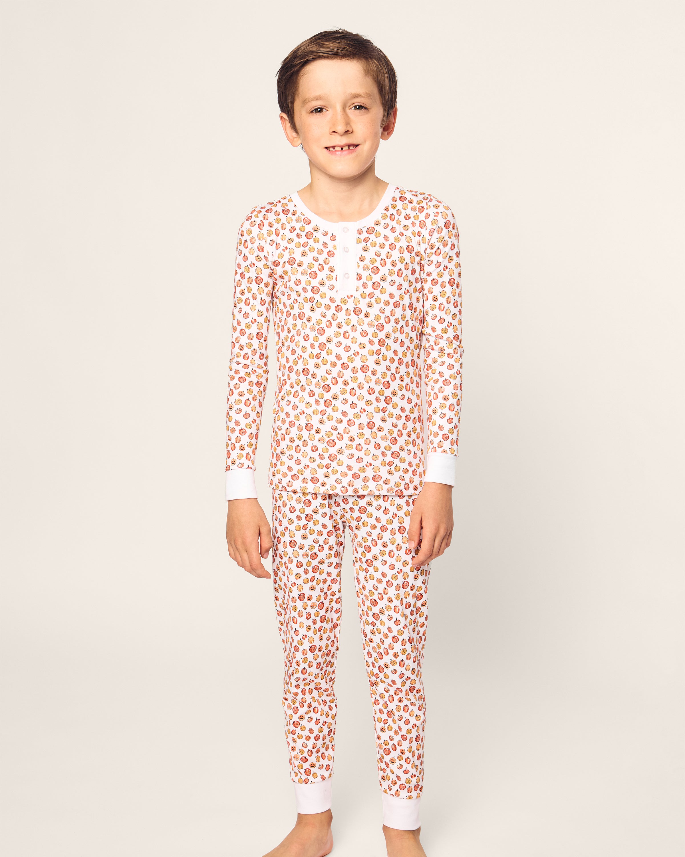 A child models Petite Plumes Kids Pima Snug Fit Pajama Set in Pumpkin Patch. Standing barefoot against a light-colored background, they smile at the camera in cozy, soft Pima Cotton pajamas featuring a playful pumpkin print with a buttoned-up, long-sleeve top.
