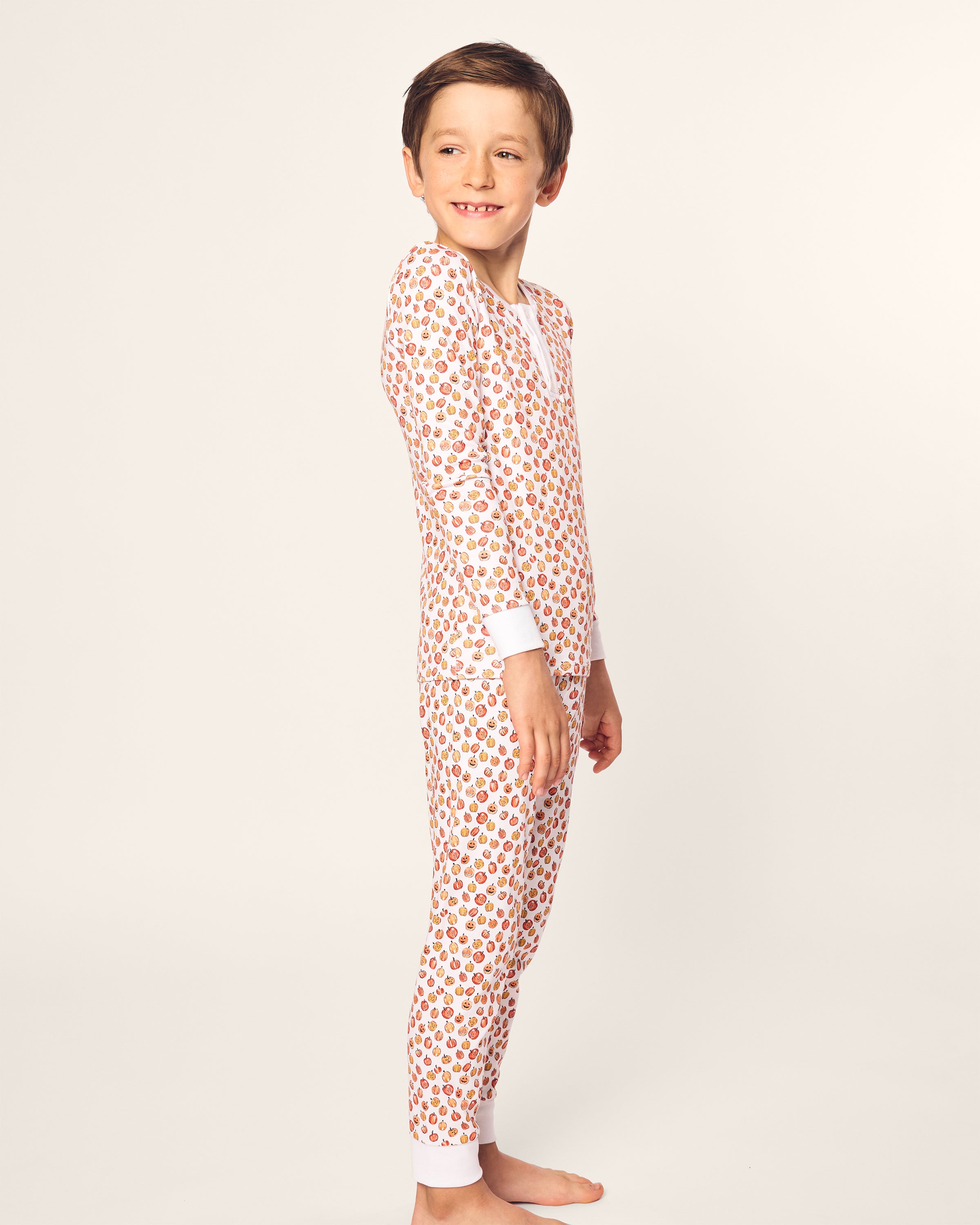 A child with short brown hair smiles while standing sideways, wearing Petite Plumes Kids Pima Snug Fit Pajama Set in Pumpkin Patch. The pajamas feature small red and brown designs on a light background, adding to the cozy vibe as the child stands barefoot against an off-white backdrop.