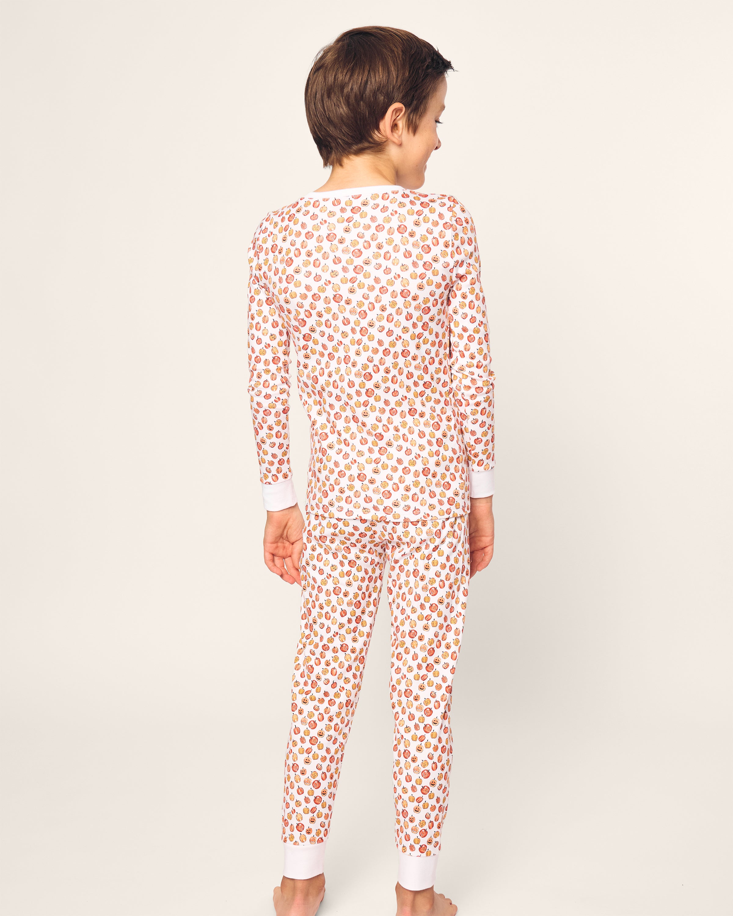 A child with short brown hair, seen from the back, wears Petite Plumes Kids Pima Snug Fit Pajama Set in Pumpkin Patch—a playful pattern on a light background. Made from soft Pima Cotton, these comfy pajamas keep them barefoot and ready for dreamland adventures.