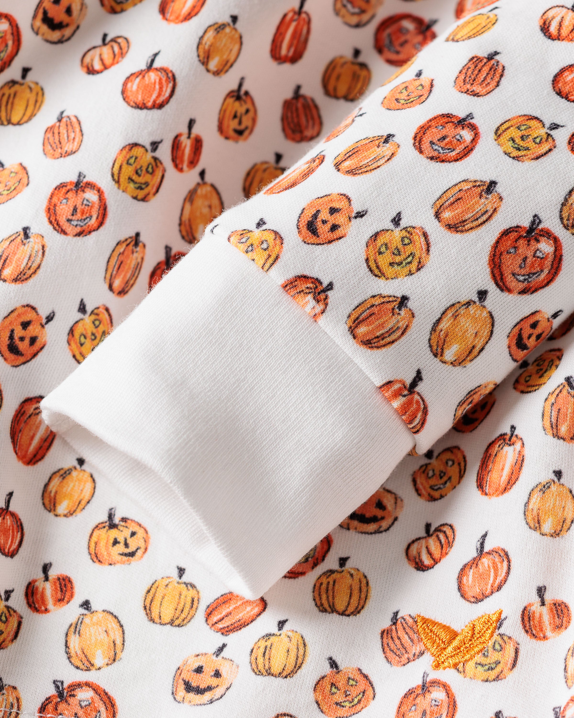 A close-up of Petite Plumes Kids Pima Snug Fit Pajama Set in Pumpkin Patch reveals a white Pima Cotton fabric with festive orange pumpkins and jack-o-lanterns. The white cuff highlights the playful autumn-themed pattern, ideal for kids sleepwear or Halloween pajamas.