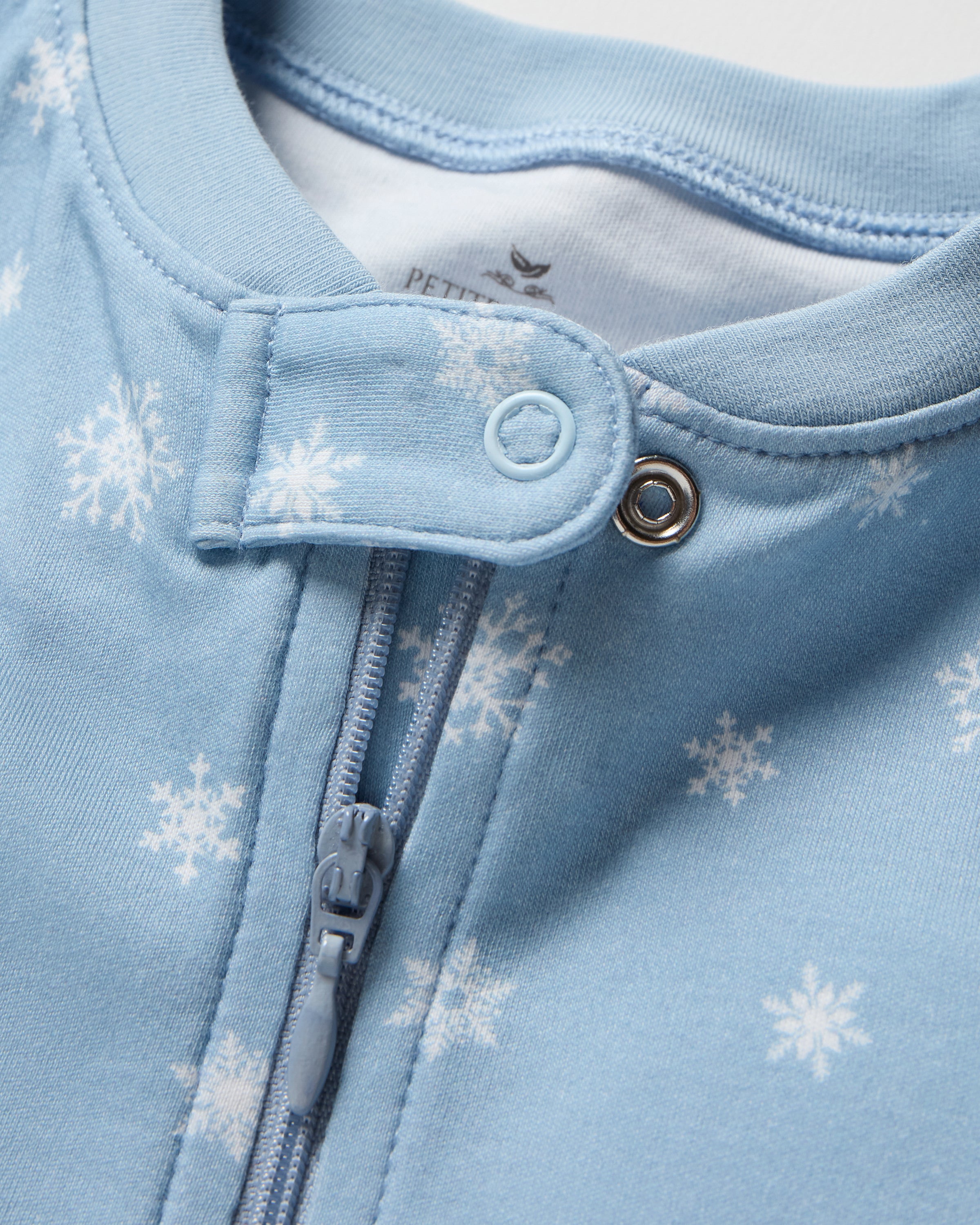 The Baby's Pima Romper in Periwinkle Wonderland is a light blue onesie with long sleeves, covered feet, white snowflake pattern, diagonal zipper from neckline to left ankle, and a small fabric snap at the neck.