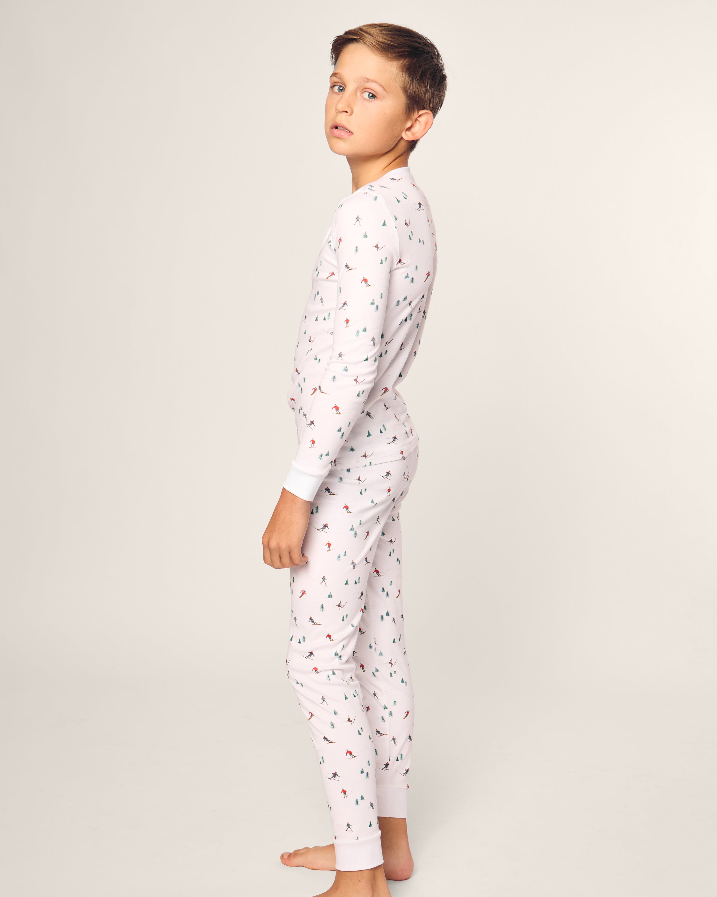 A young boy in a Petite Plume Kids Pima Snug Fit Pajama Set, Après Ski design, with long sleeves and colorful illustrations, stands barefoot on a plain backdrop, glancing over his shoulder at the camera.
