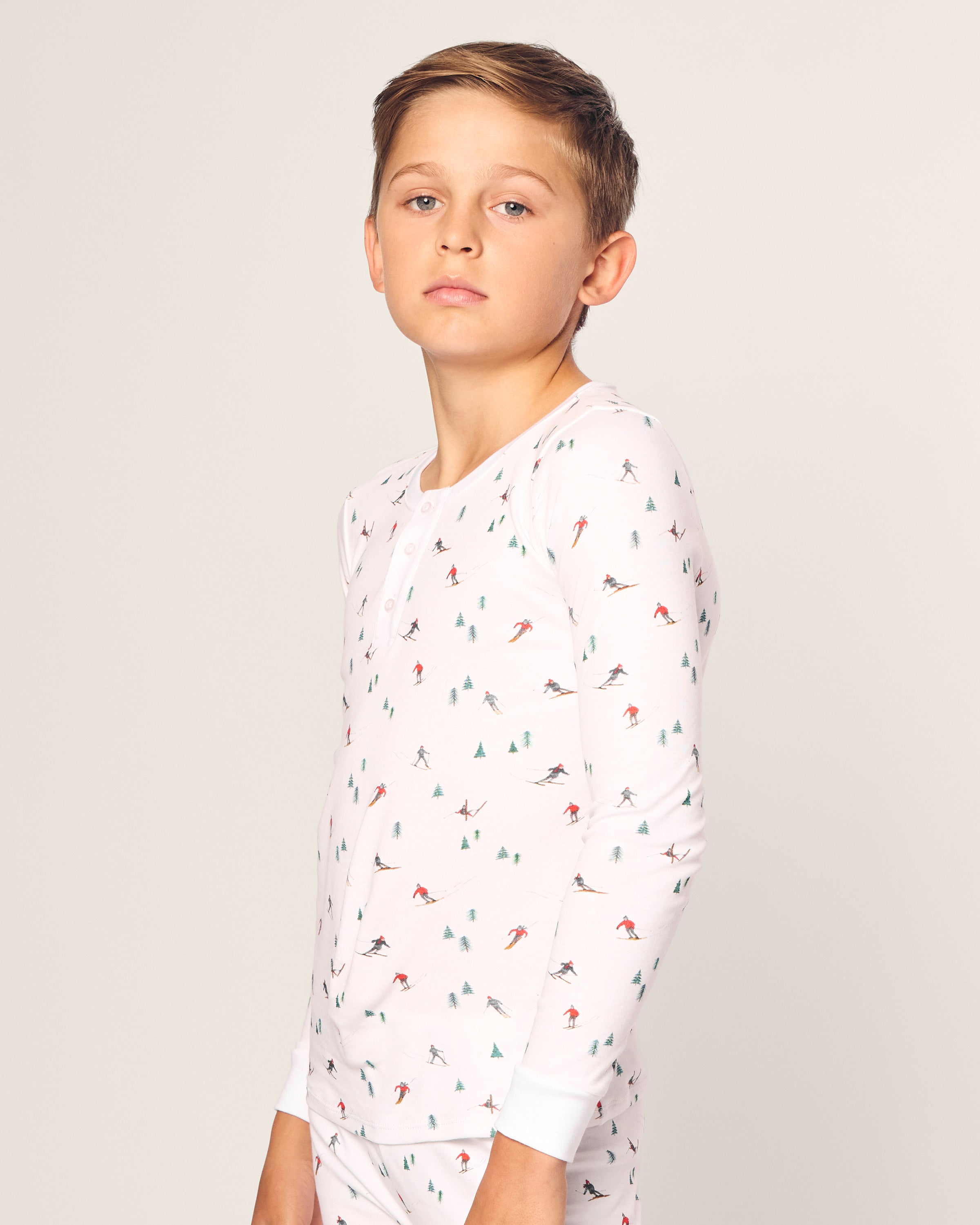 A young boy models the Kids Pima Snug Fit Pajama Set in Après Ski by Petite Plume, featuring soft white Pima cotton with skiing and tree patterns. He stands against a plain background, gazing neutrally at the camera.