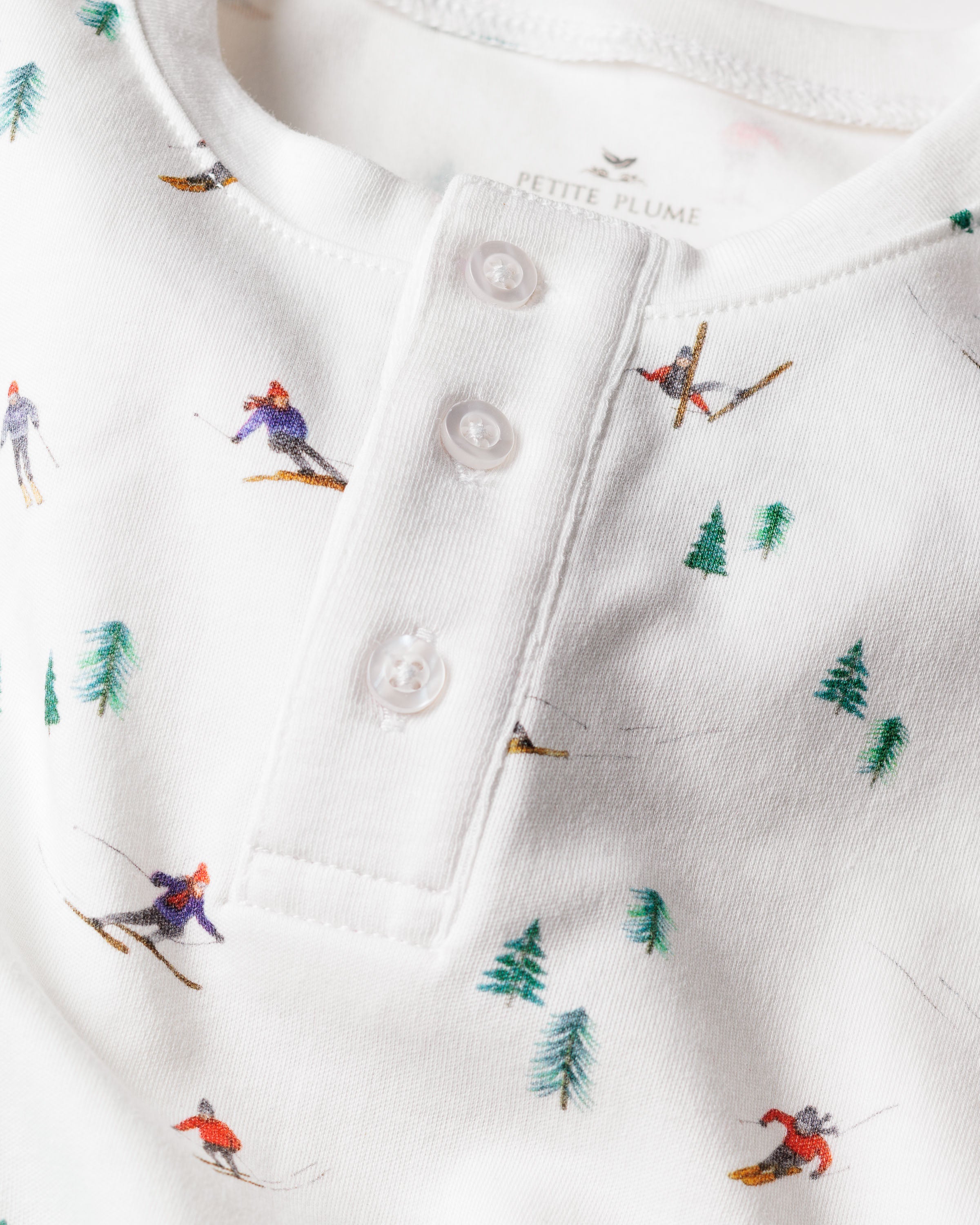A close-up of Petite Plumes Kids Pima Snug Fit Pajama Set in Après Ski shows a white long-sleeve top adorned with small skiers and green pine trees, featuring a soft texture and a buttoned placket with three white buttons.